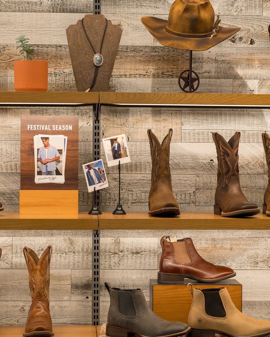 Cowboy boots displayed on a wooden shelf, with a vintage cowboy hat, a necklace, and small photos of a person wearing western clothing, along with a potted plant and a poster that reads 'Festival Season'.