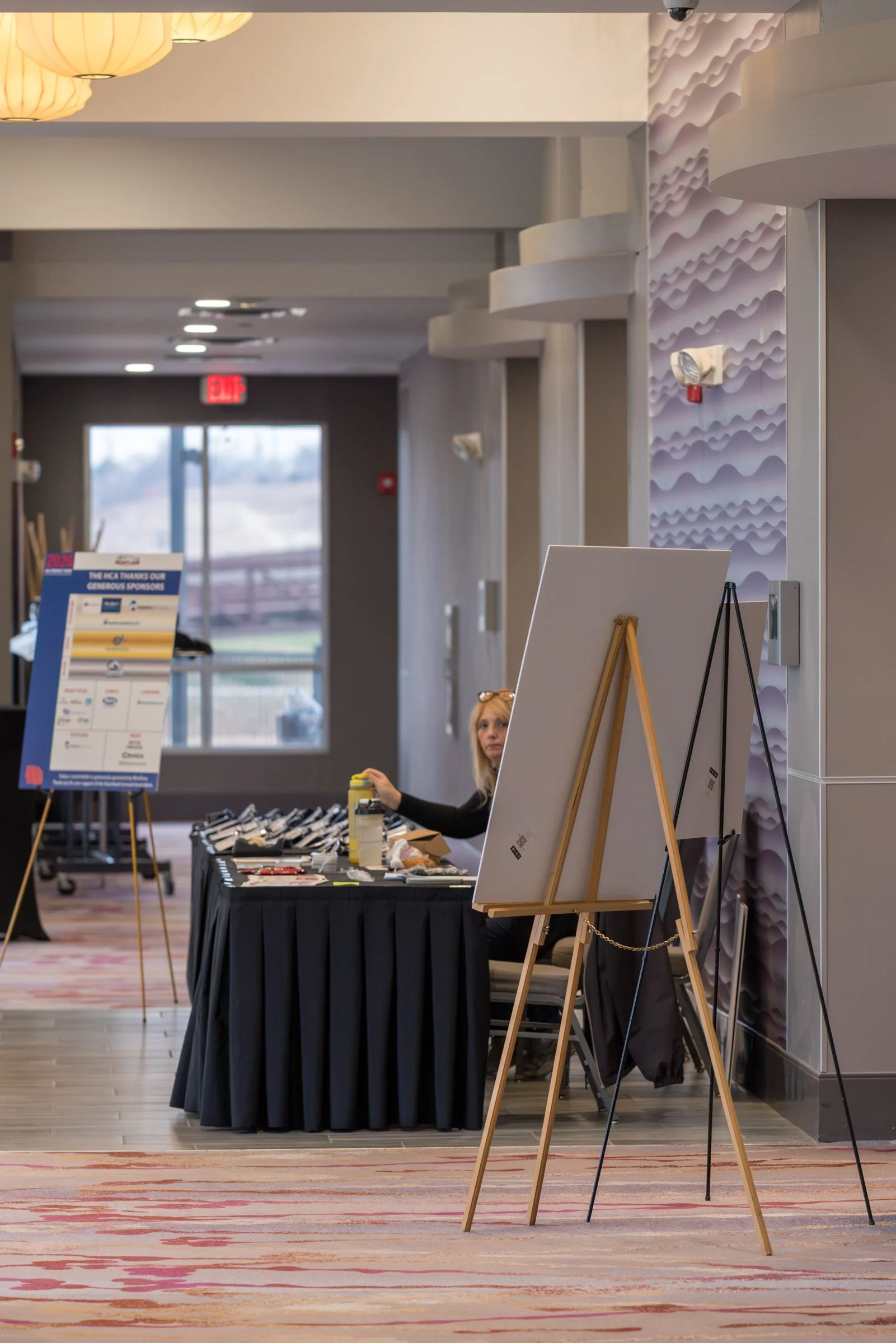 Interior of a conference or event space with a table displaying items, a woman seated behind the table, and various signs and display boards.