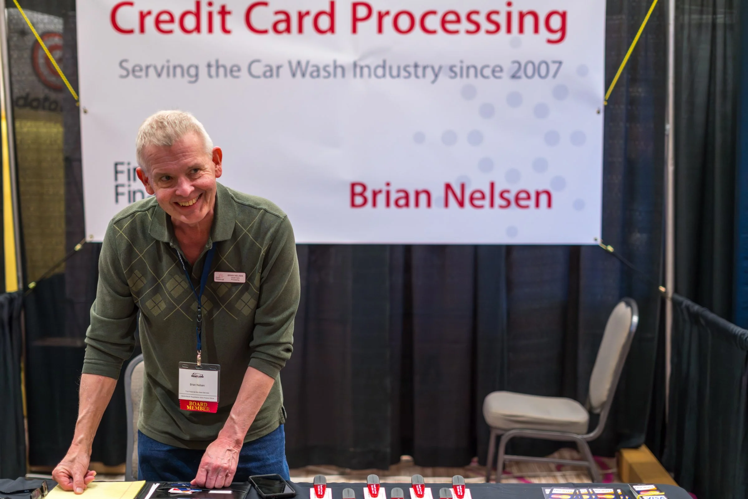 A smiling man with gray hair and a green sweater standing at a booth with a banner that reads 'Credit Card Processing, Serving the Car Wash Industry since 2007, Brian Nelsen.' He is wearing a name badge and a red 'Board Member' tag, engaging with ele