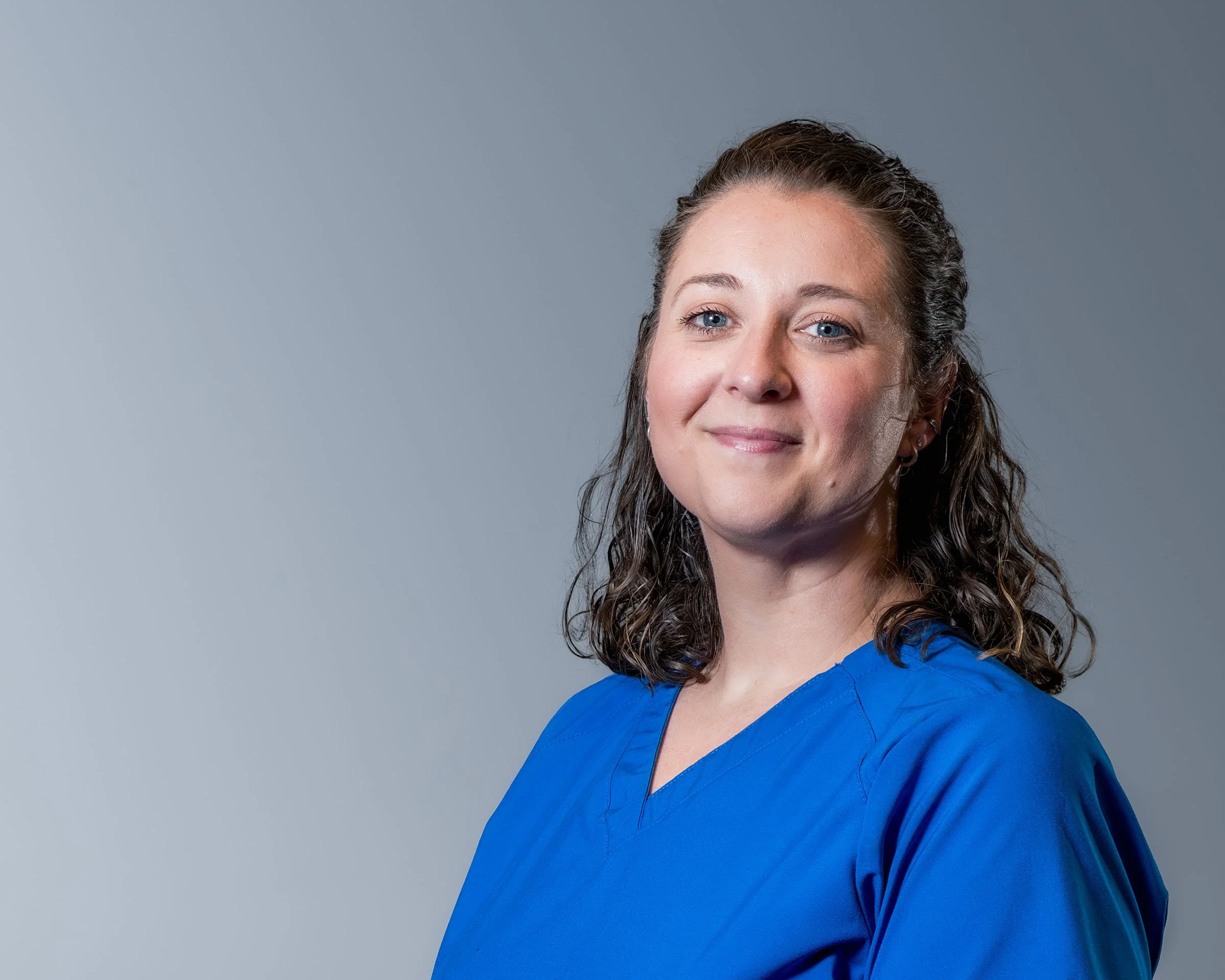 A woman with wet, wavy hair and blue eyes wearing a blue medical scrub top, standing against a gray background.