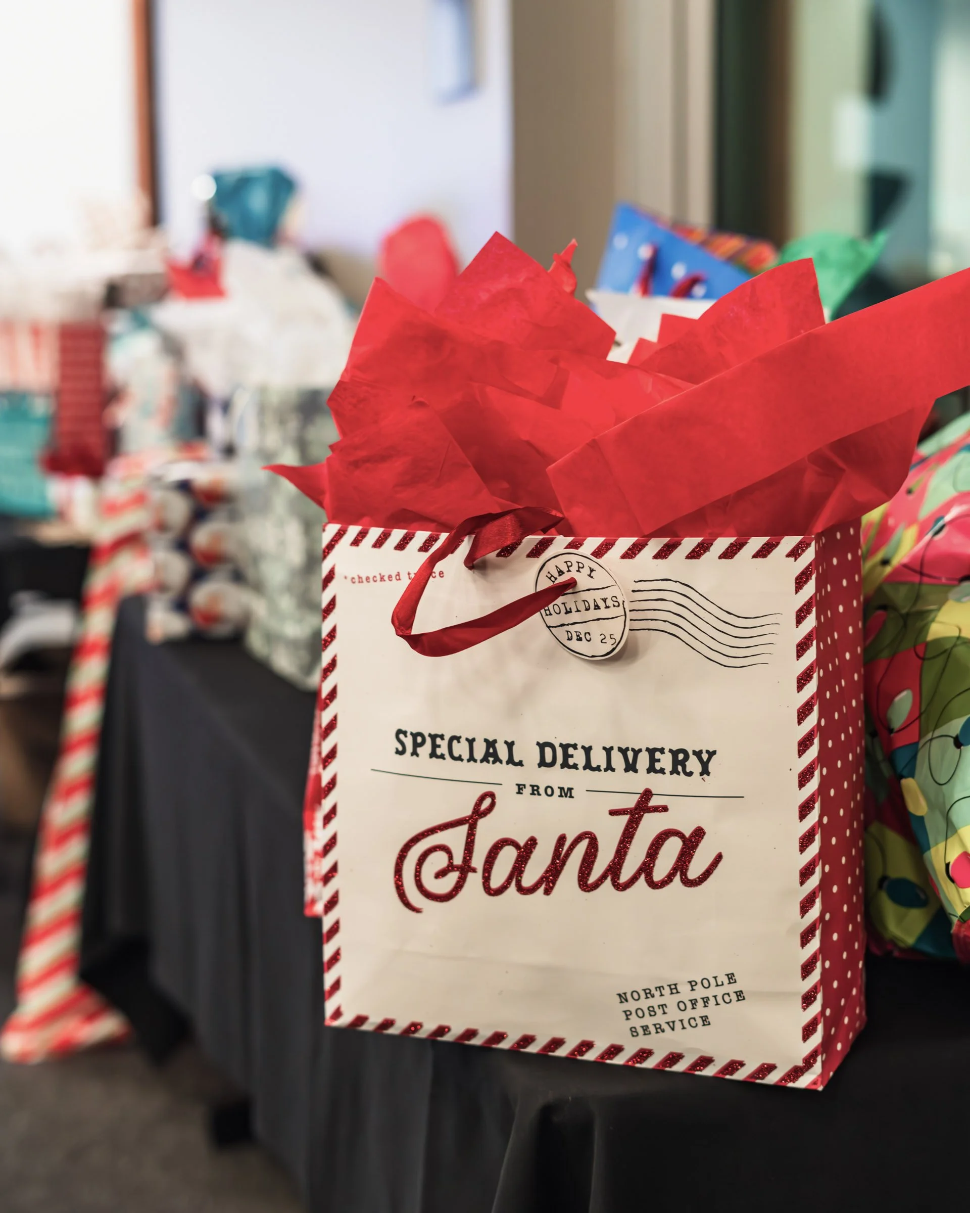 A festive holiday gift bag with red tissue paper on a black surface, with blurred holiday decorations and presents in the background.