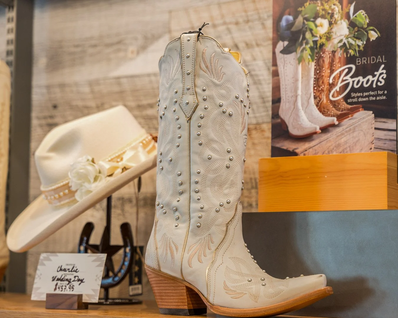 White cowboy boot decorated with pearl-like studs and embroidery, displayed on a shelf with a sign indicating it is a Charlie Valentine Valentine's Day boot priced at $457.95, and a white hat with a floral decoration in the background