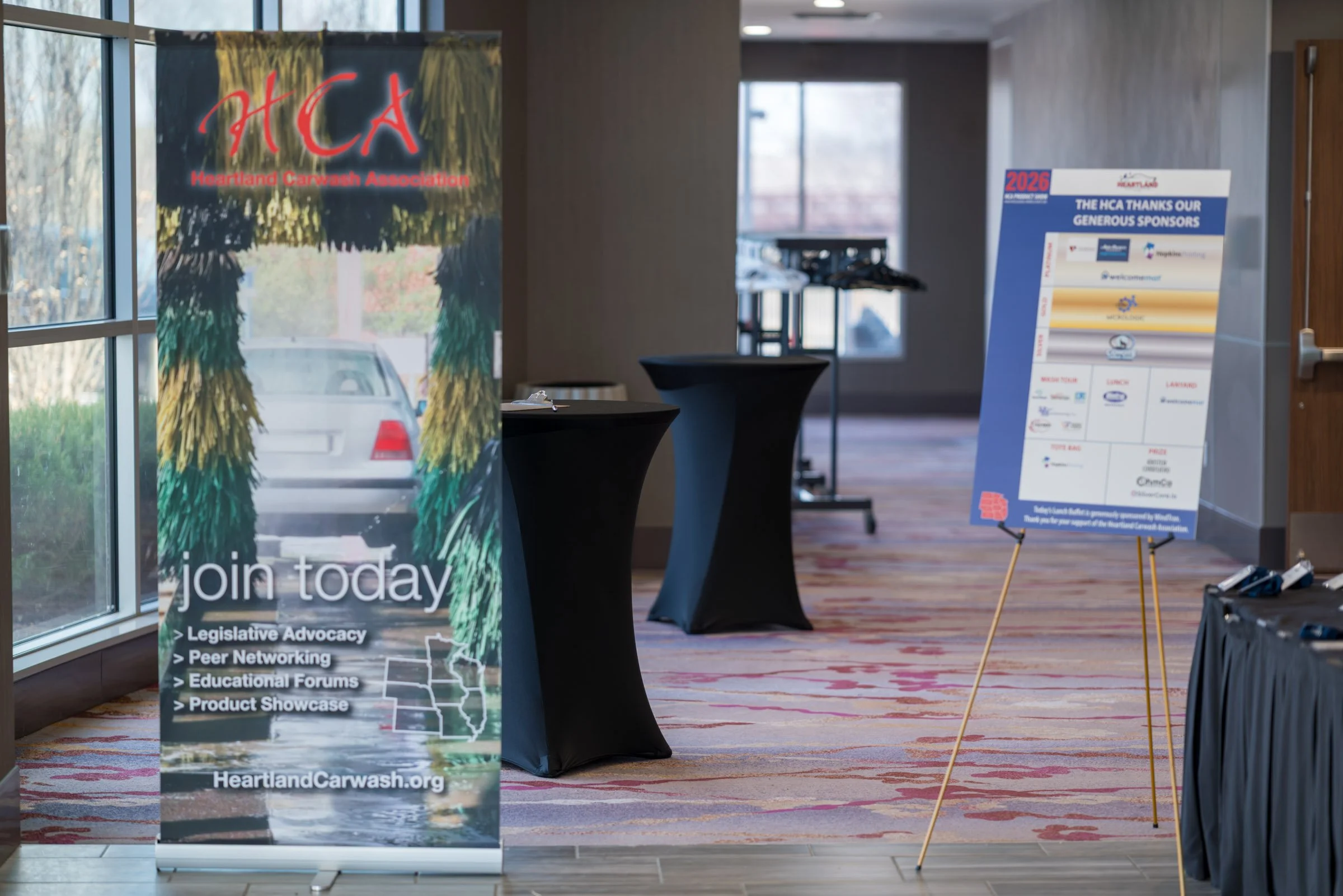 Indoor conference or event area with banners and tables. One banner features the Heartland Carwash Association logo and promotes joining for legislative advocacy, peer networking, educational forums, and product showcases. Another sign lists sponsors