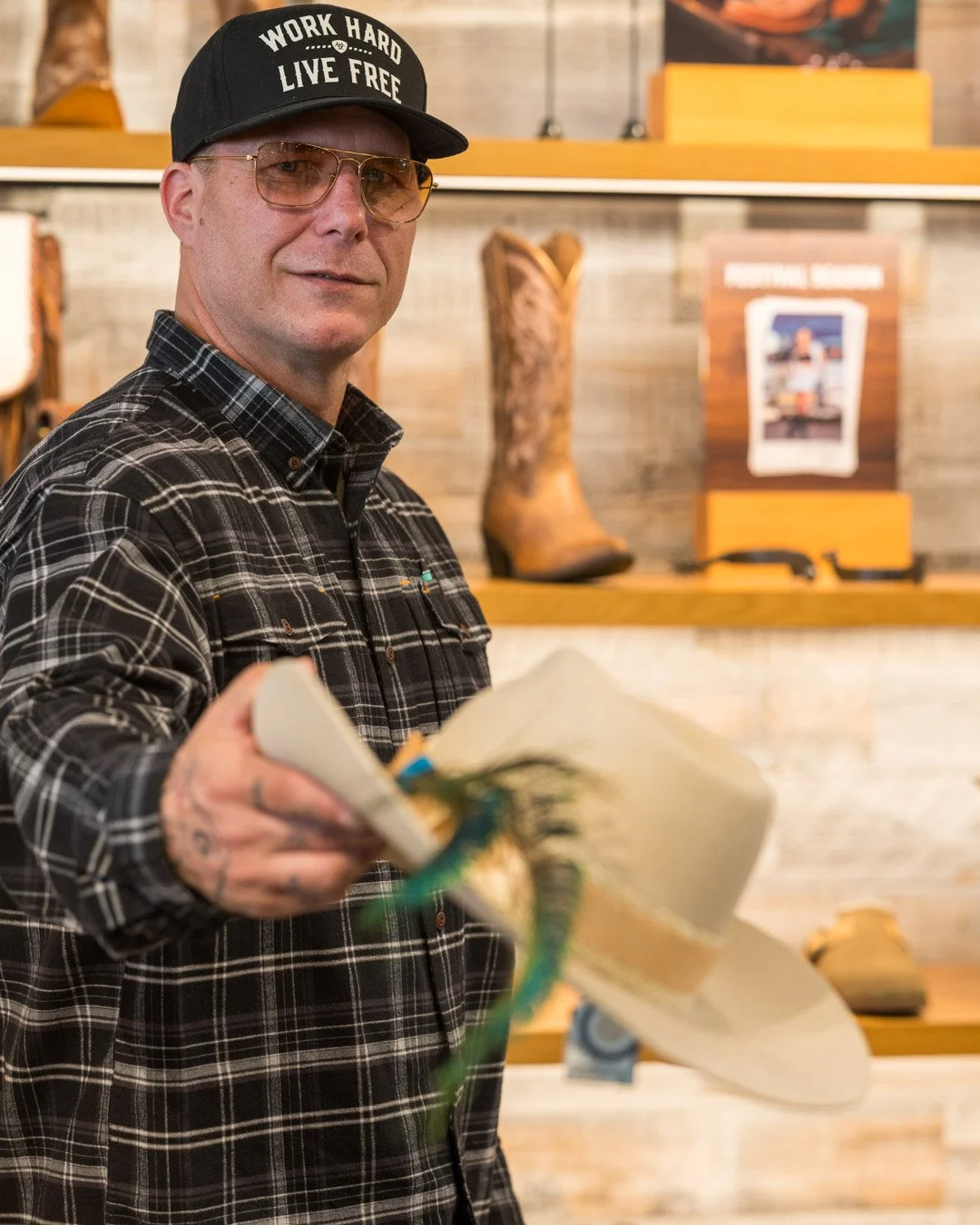 A man wearing glasses, a black cap that says "Work Hard Live Free," and a black plaid shirt, holding a white cowboy hat with a green band in a store with Western boots and rustic decor in the background.