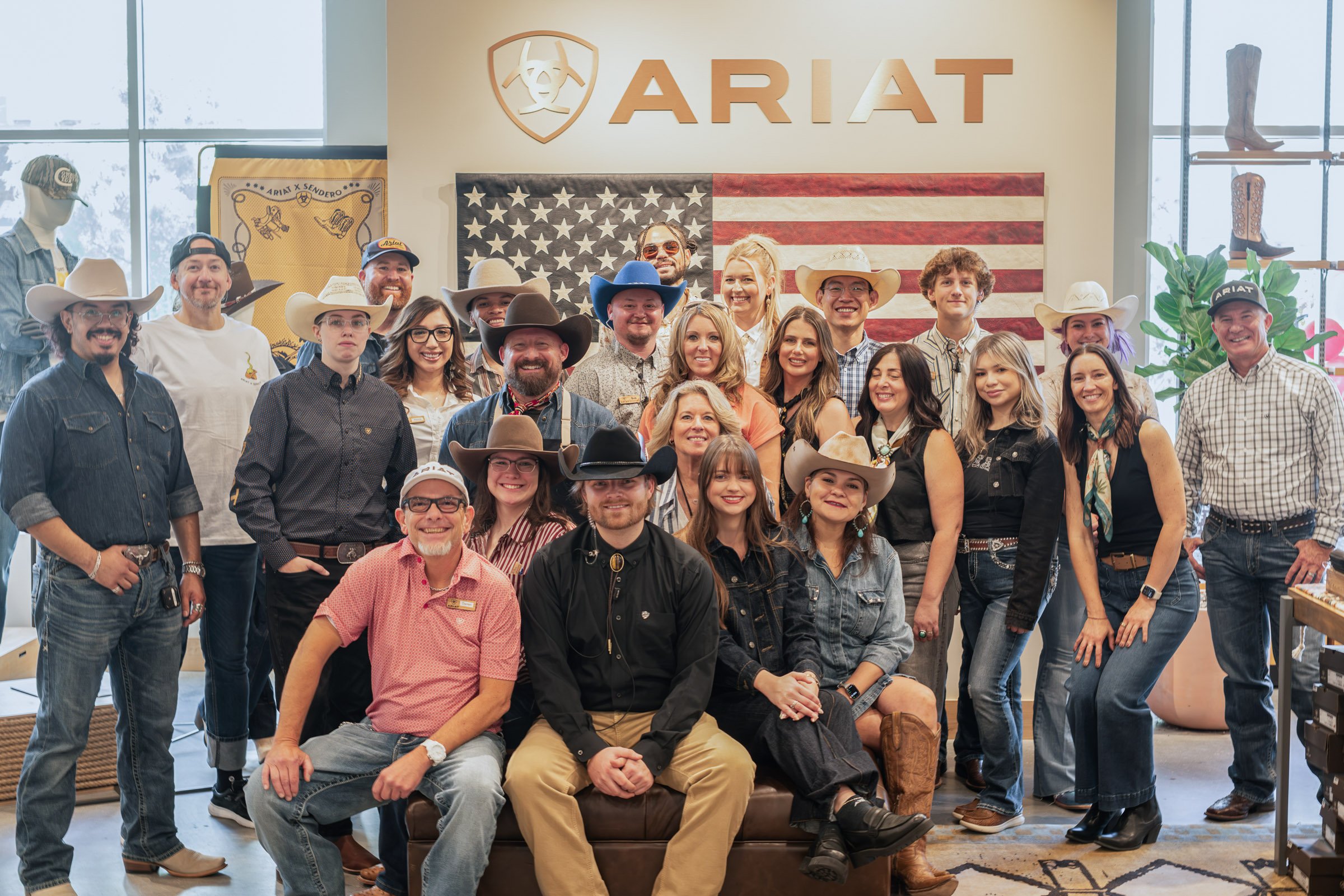 Group of people gathered indoors, some wearing cowboy hats, in front of a wall with the ARIAT logo and an American flag, with some cowboy boots displayed on shelves.