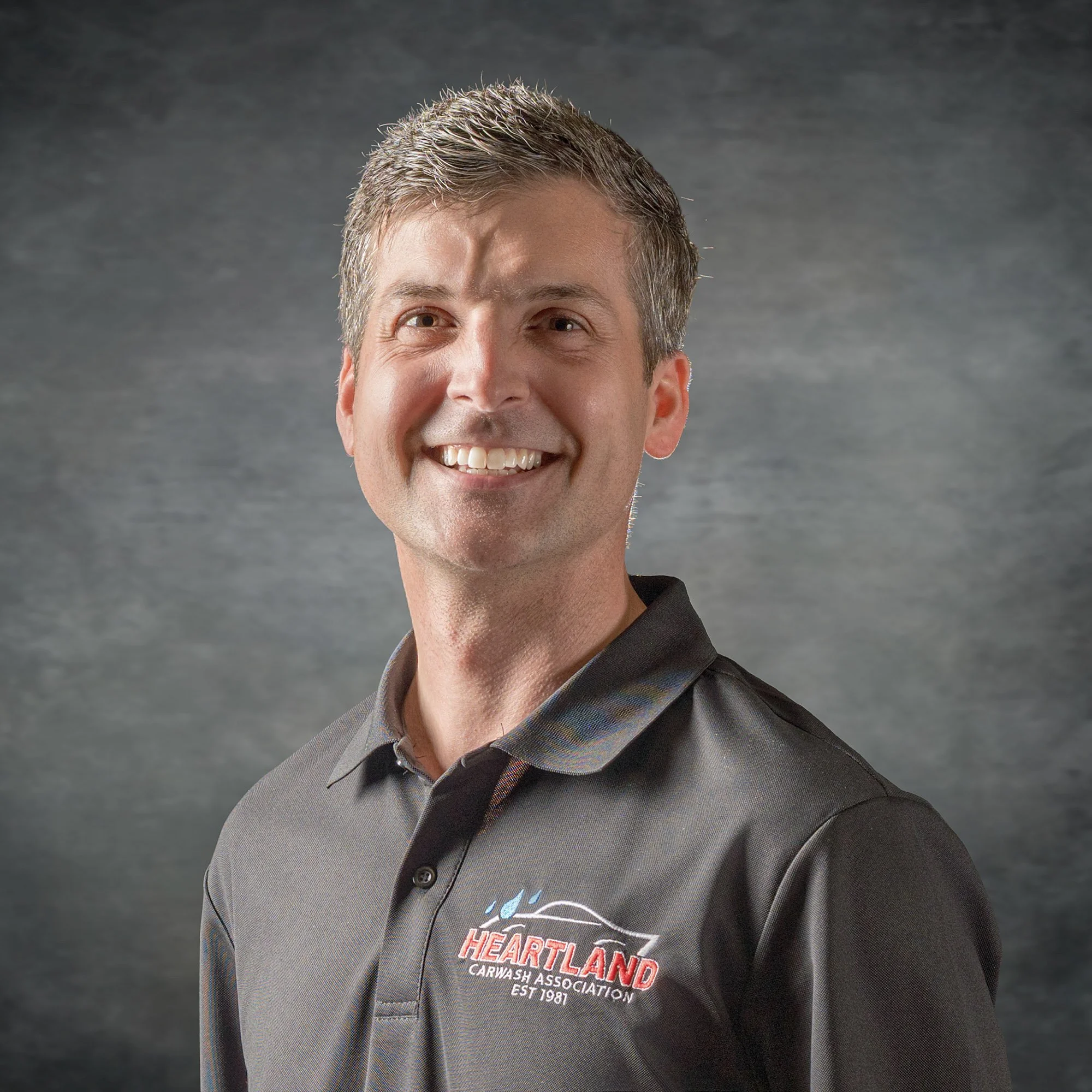 Close-up of a smiling man wearing a gray jacket with an embroidered logo reading "Heartland Carwash" on the chest, set against a dark, textured background.
