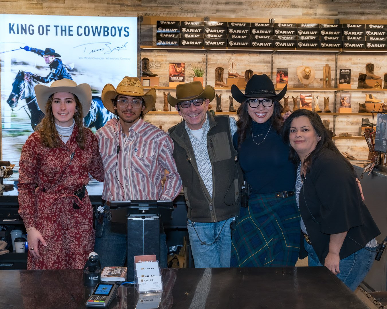 Five people in Western attire standing inside a cowboy boot shop, with a large digital screen behind them displaying a rodeo image and text that reads 'King of the Cowboys.' They are smiling and posing for the photo, with shelves of cowboy boots and 