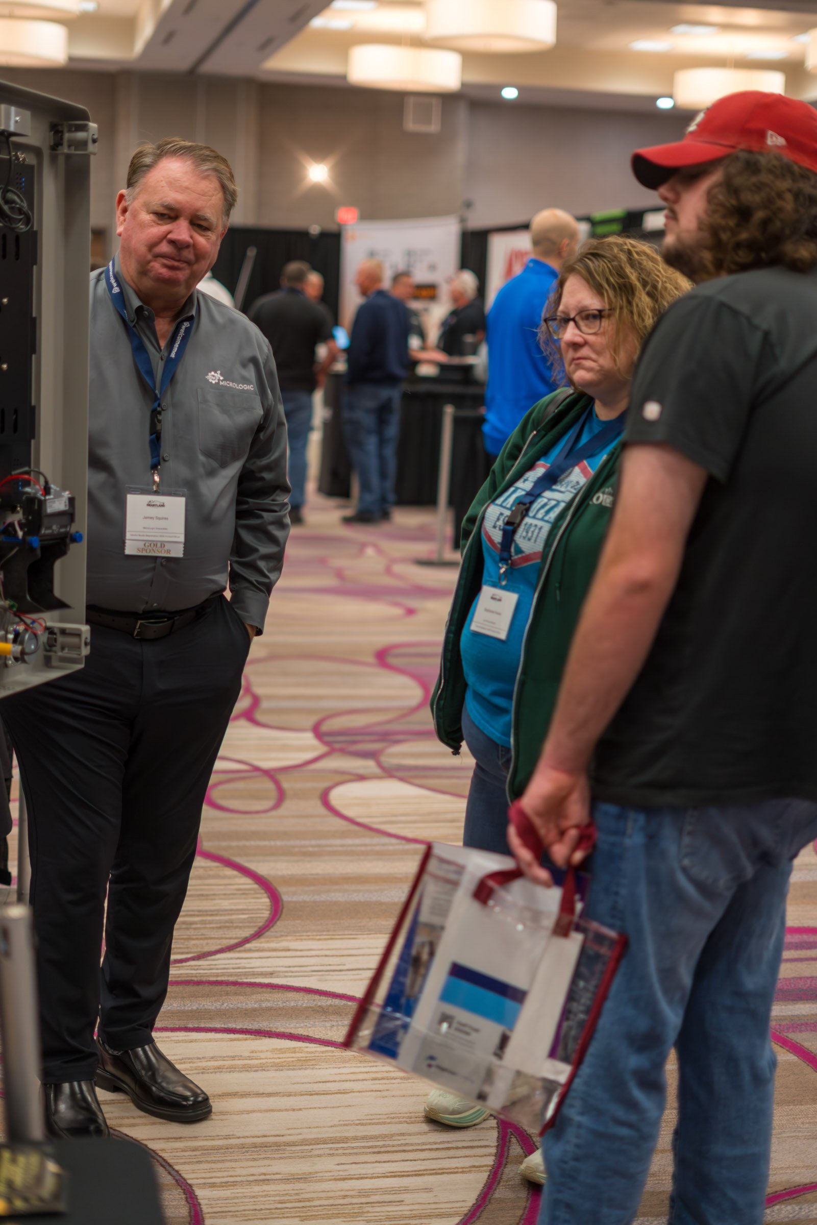 Three people are having a conversation at a trade show or conference. Two women and one man stand close together, with the man on the left side of the image near what appears to be a display or demonstration. The background shows other attendees and 