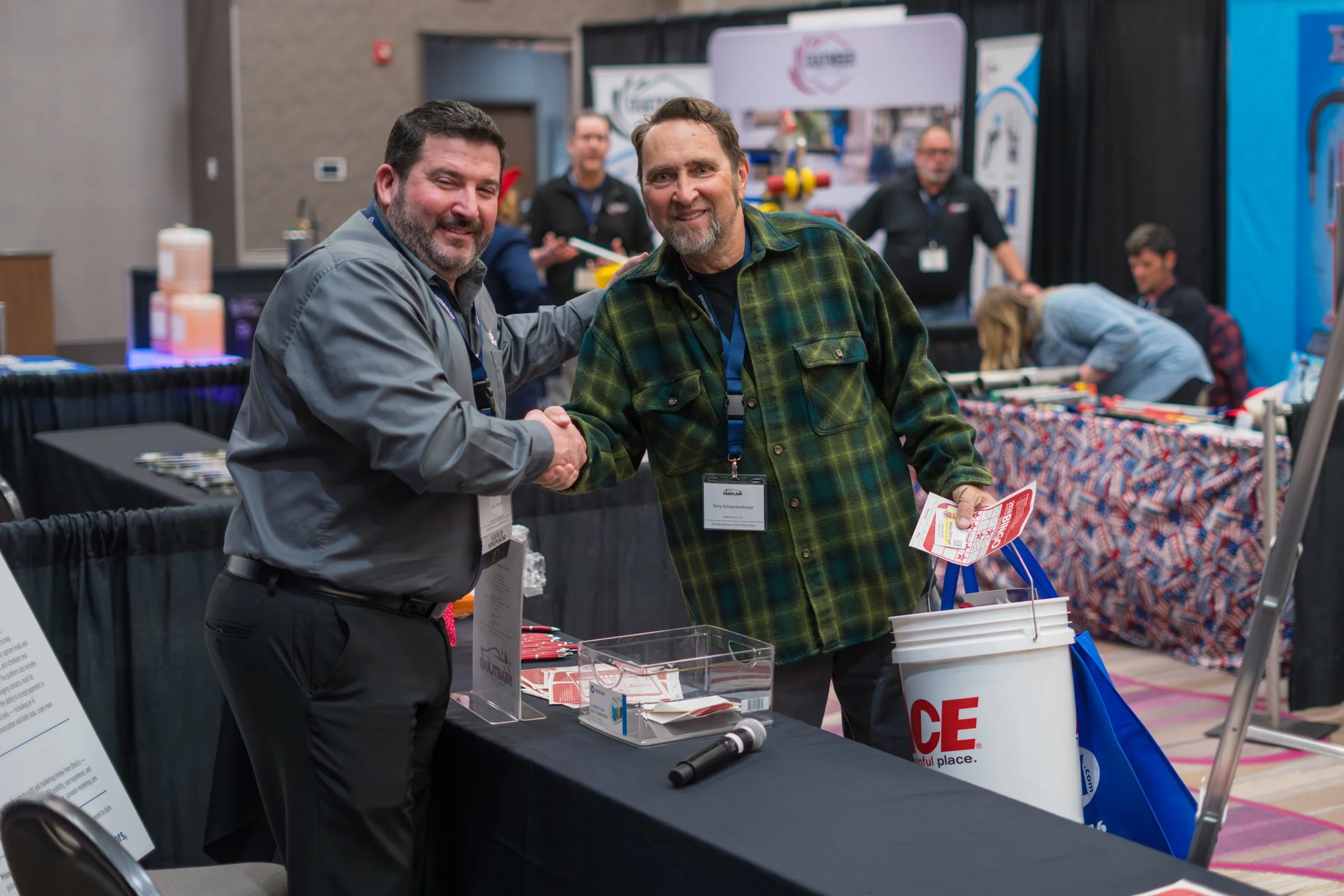 Two men shake hands at a conference or trade show, with several people and display booths in the background.