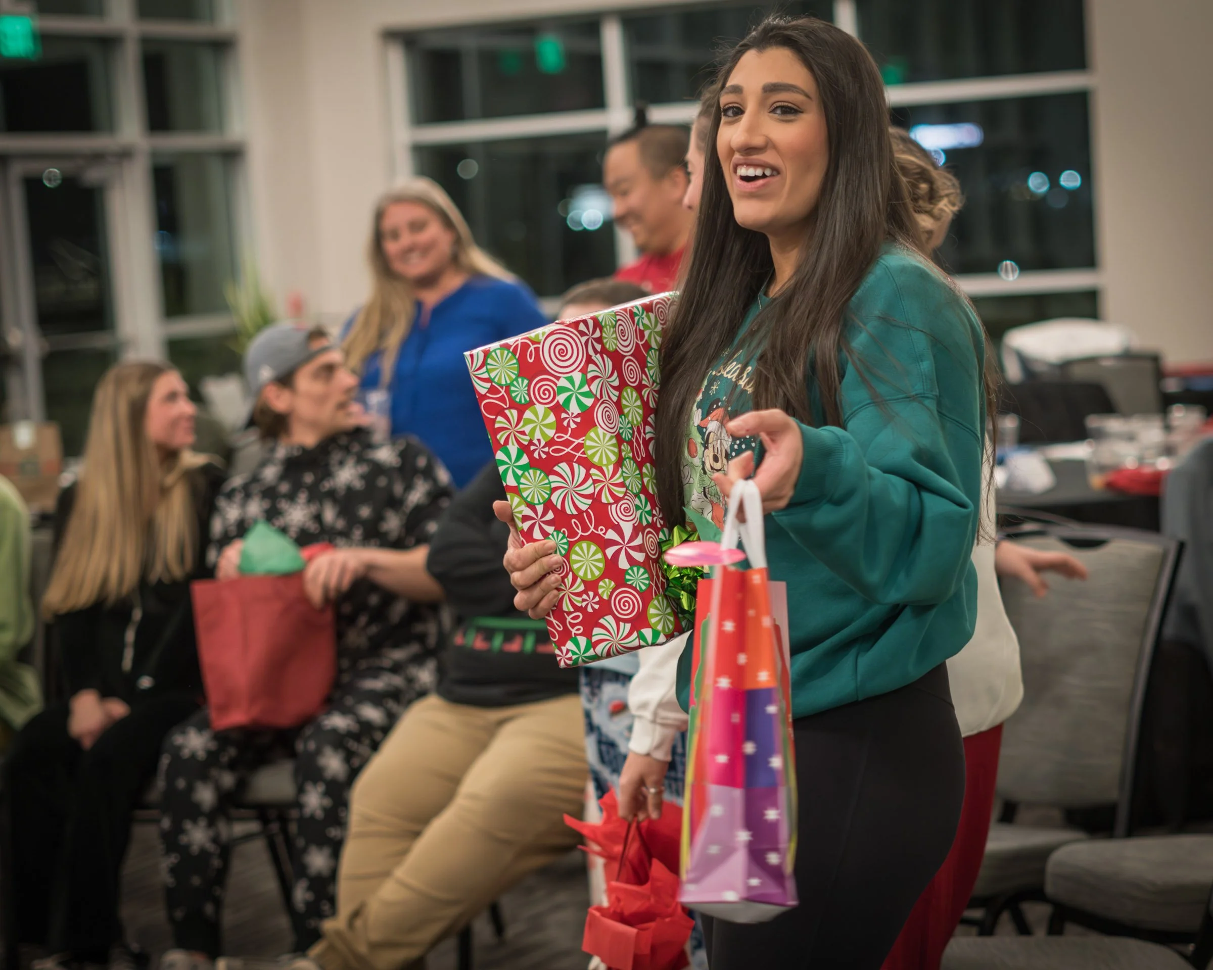 A group of people sitting and standing at a holiday party, one woman in a teal sweater holding a large present with holiday-themed wrapping, smiling, with other gathered adults in the background.
