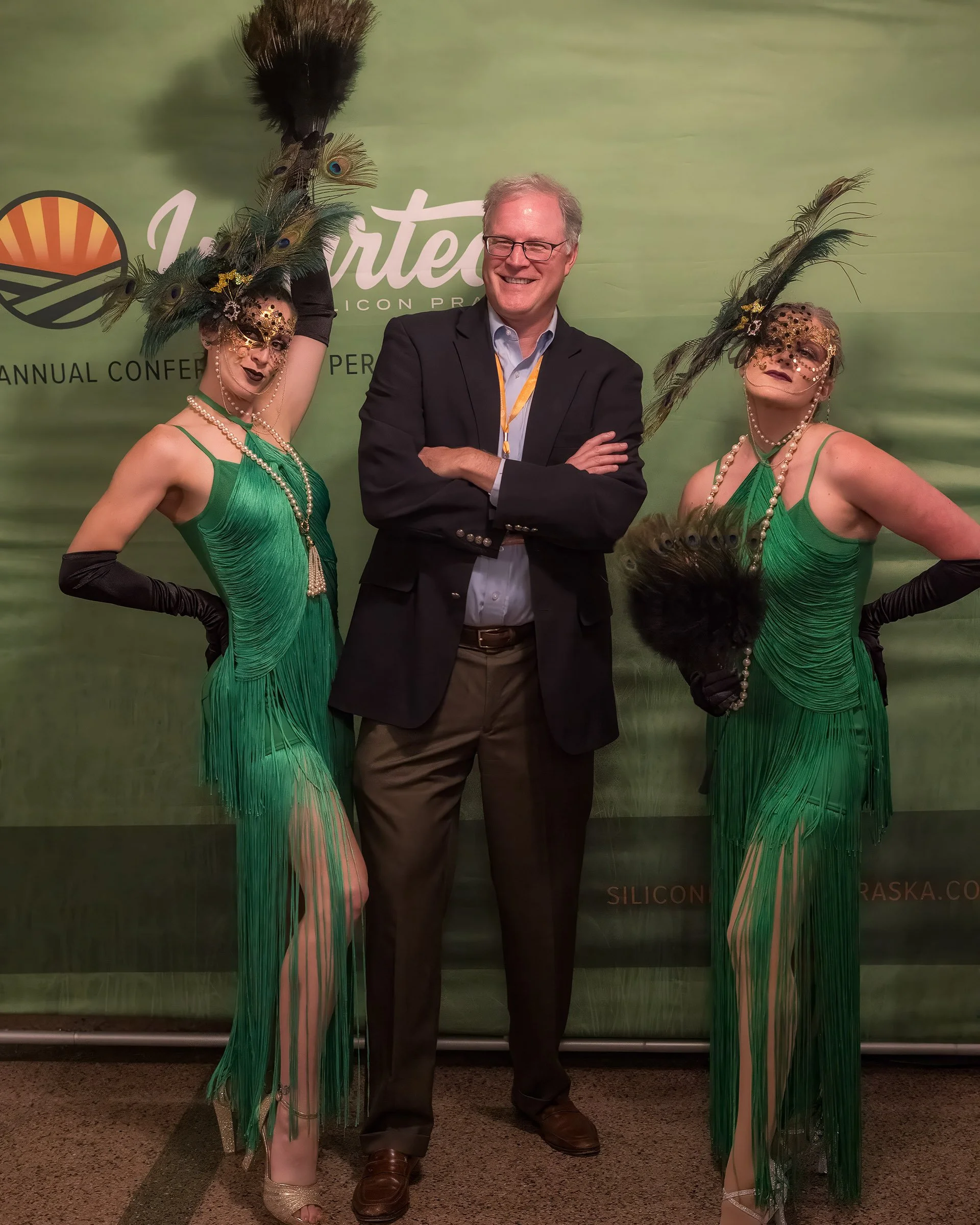 A man in a dark suit with glasses smiling, standing between two women dressed in green dresses with feathered headpieces, pearl necklaces, and gloves, posing at an event with a green backdrop.