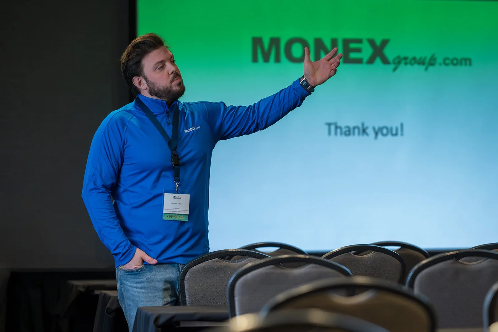 A man wearing a blue jacket and a conference badge is standing and pointing towards a presentation screen that displays the logo 'MONEGX group.com' and a 'Thank you!' message, with empty chairs in the foreground.