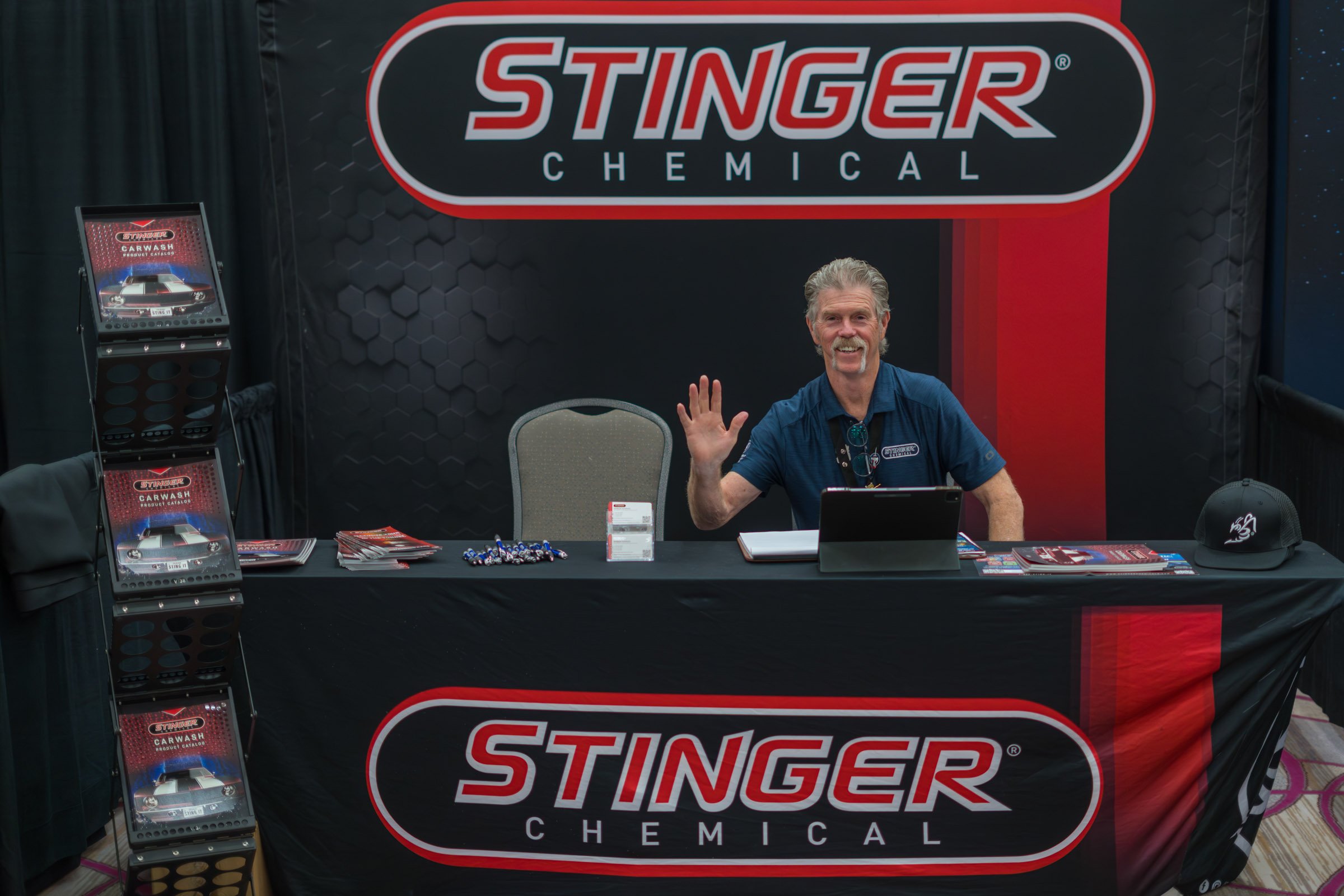 A man sitting at a table with promotional materials for Stinger Chemical, waving and smiling. The backdrop has the Stinger Chemical logo; the table has brochures, pens, and a laptop.