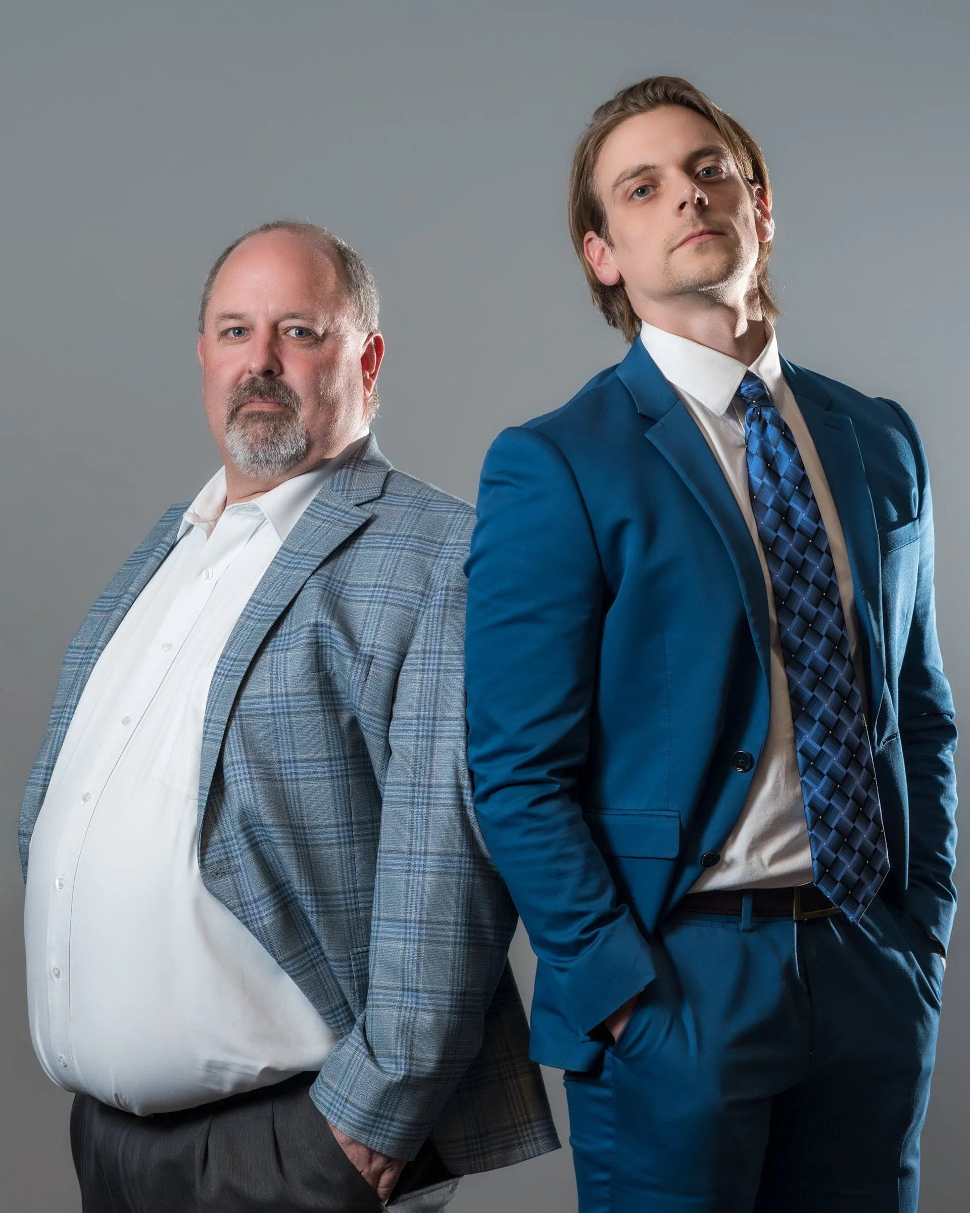 Two men dressed in business suits standing side by side against a gray background. The man on the left has a beard and is wearing a light gray checkered suit with a white shirt. The man on the right has medium-length hair and is wearing a bright blue