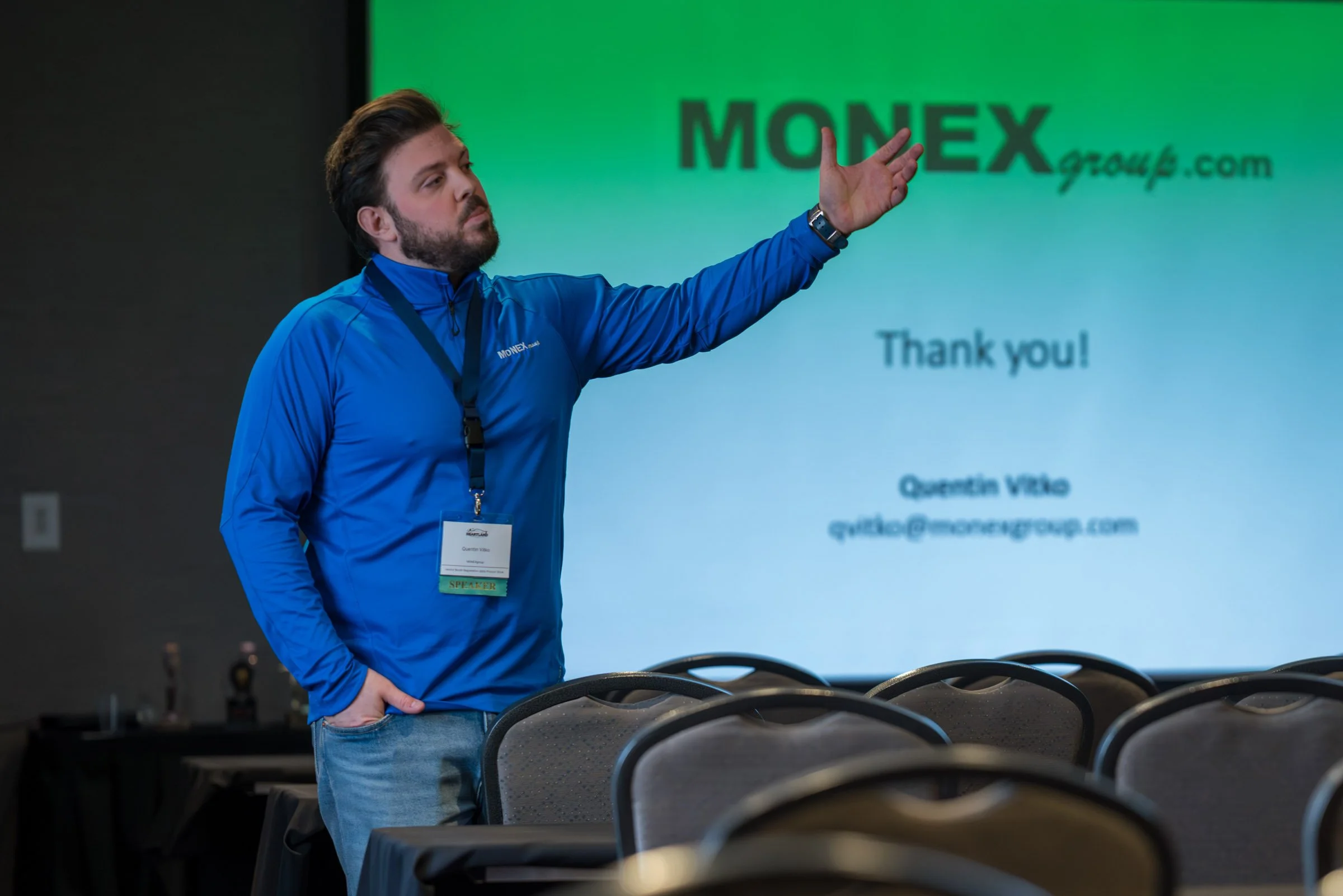 A man wearing a blue jacket and conference badge presenting in front of a screen with the logo 'MONEY group.com' and a message saying 'Thank you!'