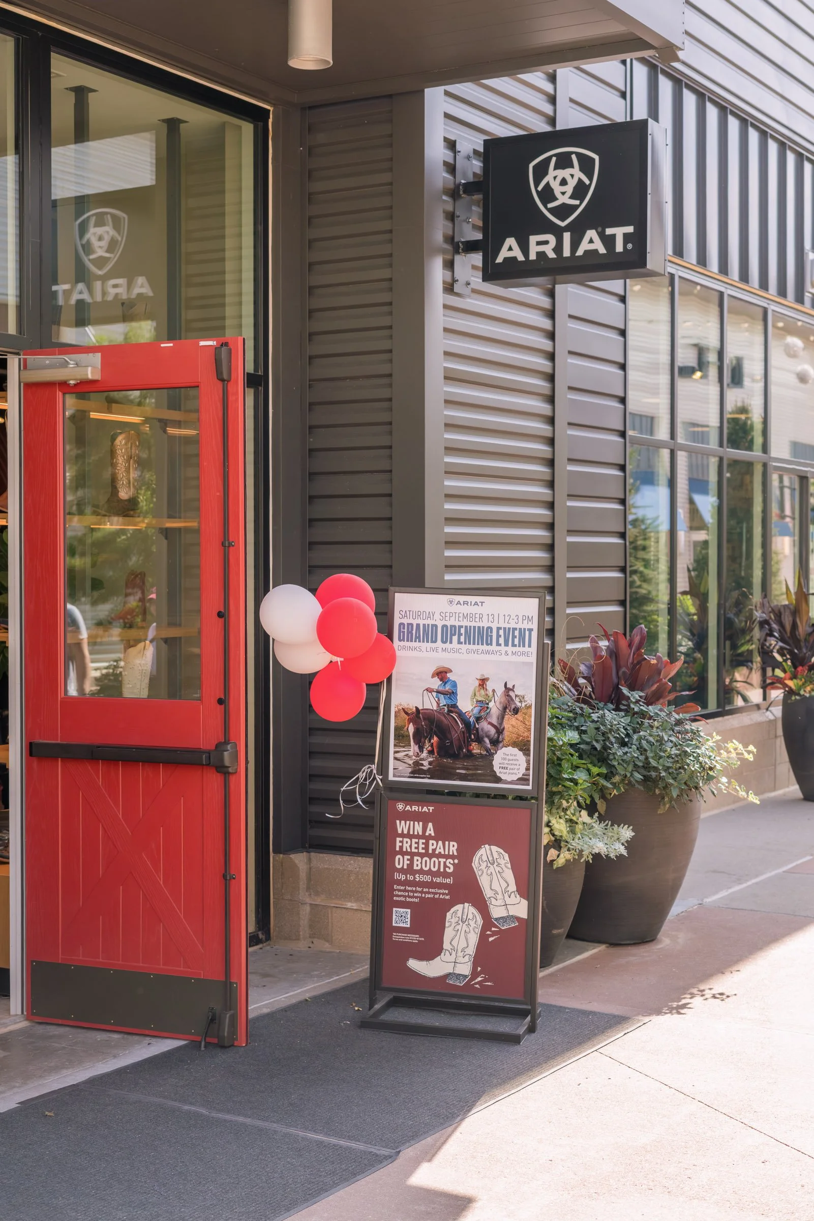 The exterior of an ARIAT store with a black sign featuring a shield logo and the brand name. There is a red door and promotional event sign with balloons and a large potted plant nearby.
