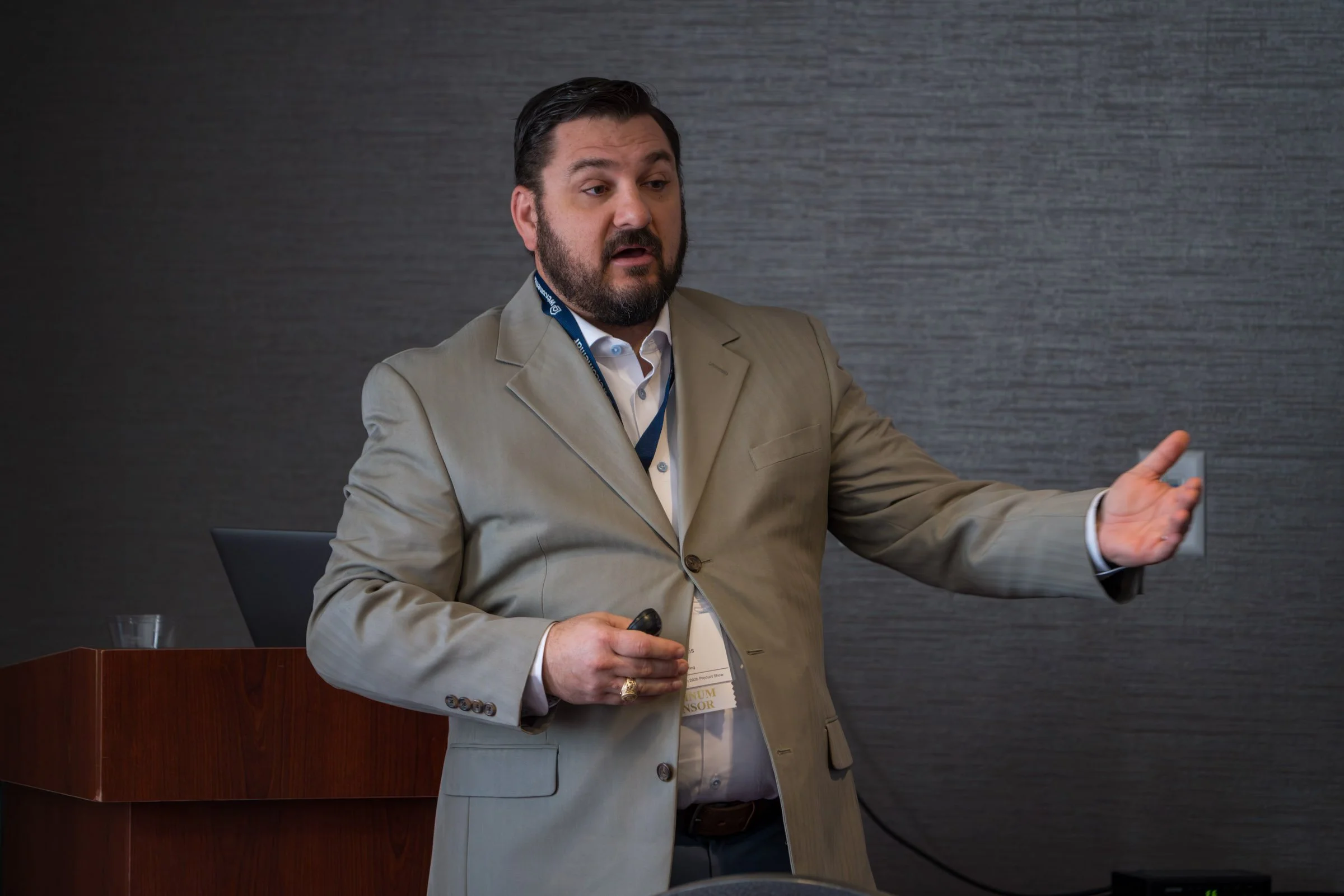 A man in a beige suit is speaking and gesturing with his right hand during a presentation or meeting, with a dark textured wall in the background.