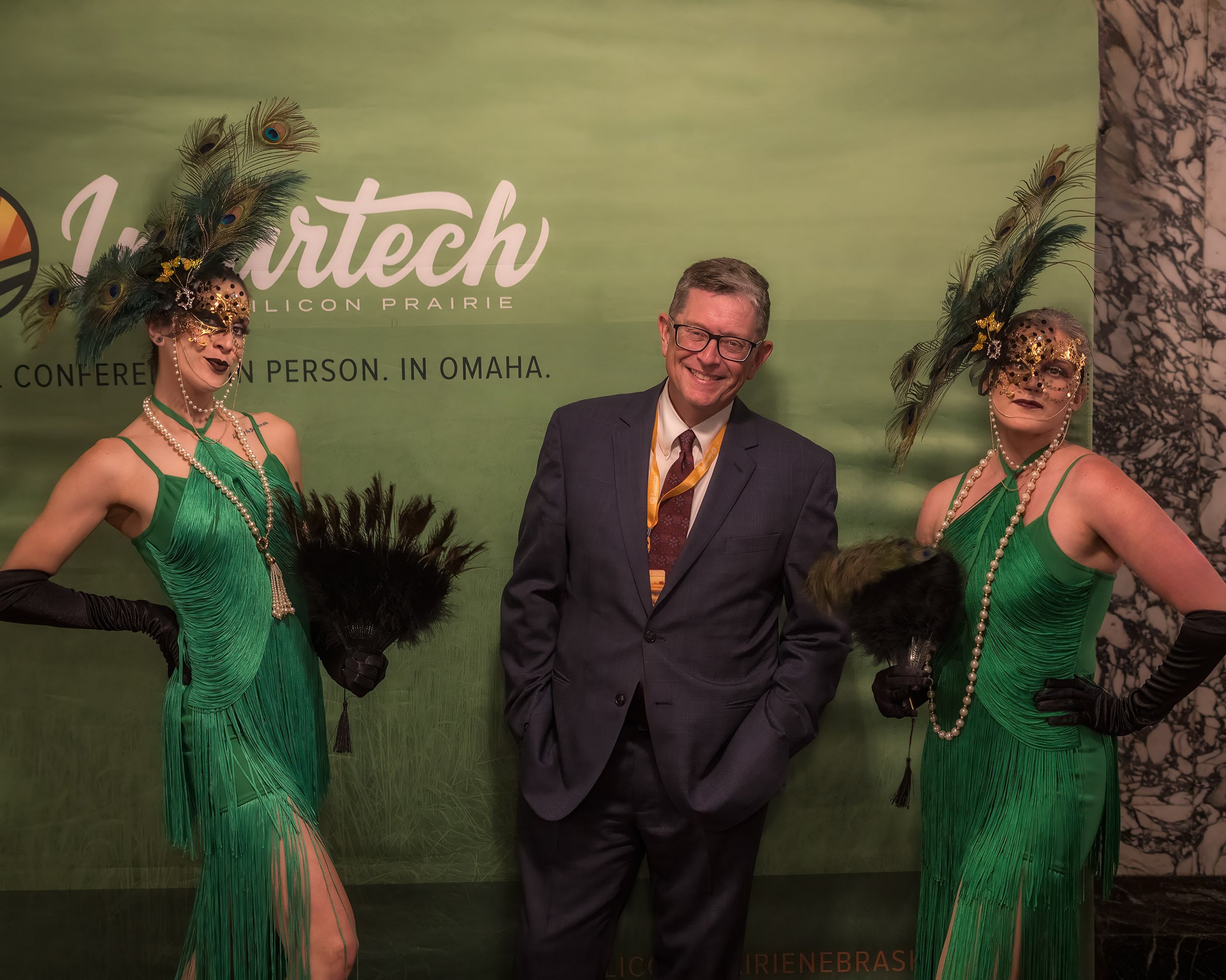 Two women dressed in green costumes with feathered headdresses and pearl jewelry, and a man in a suit, standing in front of a green backdrop with text.