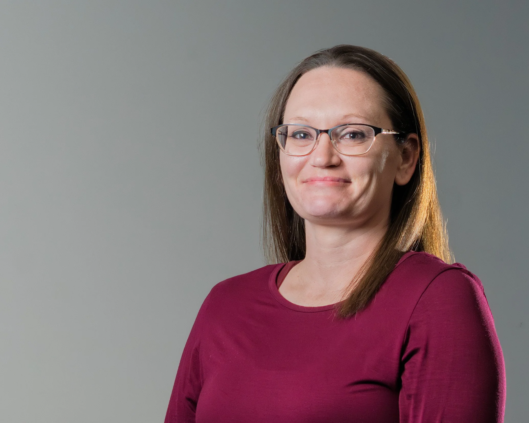 A woman with long brown hair wearing glasses and a maroon long sleeve shirt, smiling against a plain gray background.