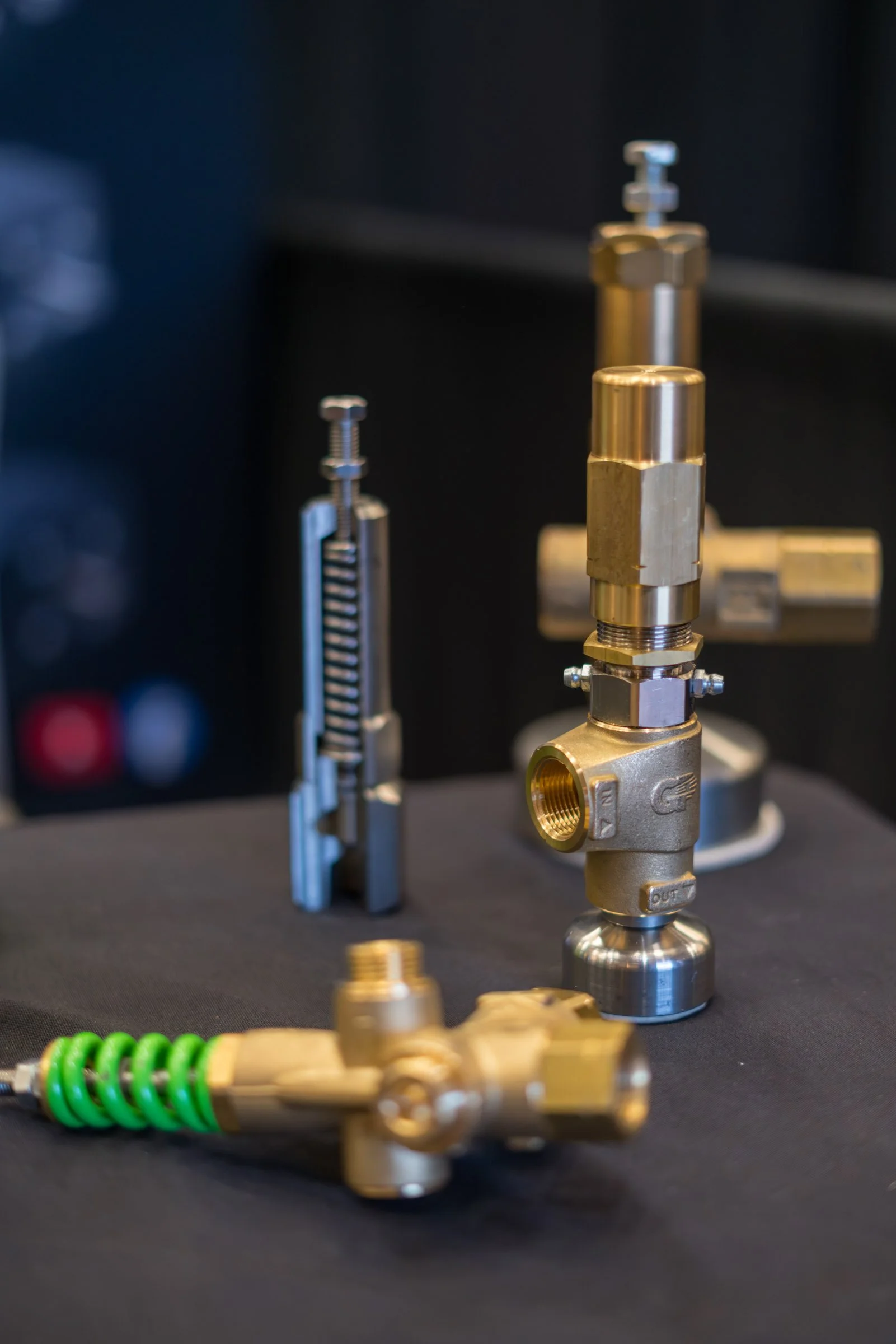 Close-up of metal industrial components, including brass and steel fittings, valves, and connectors, arranged on a black surface with a dark background.