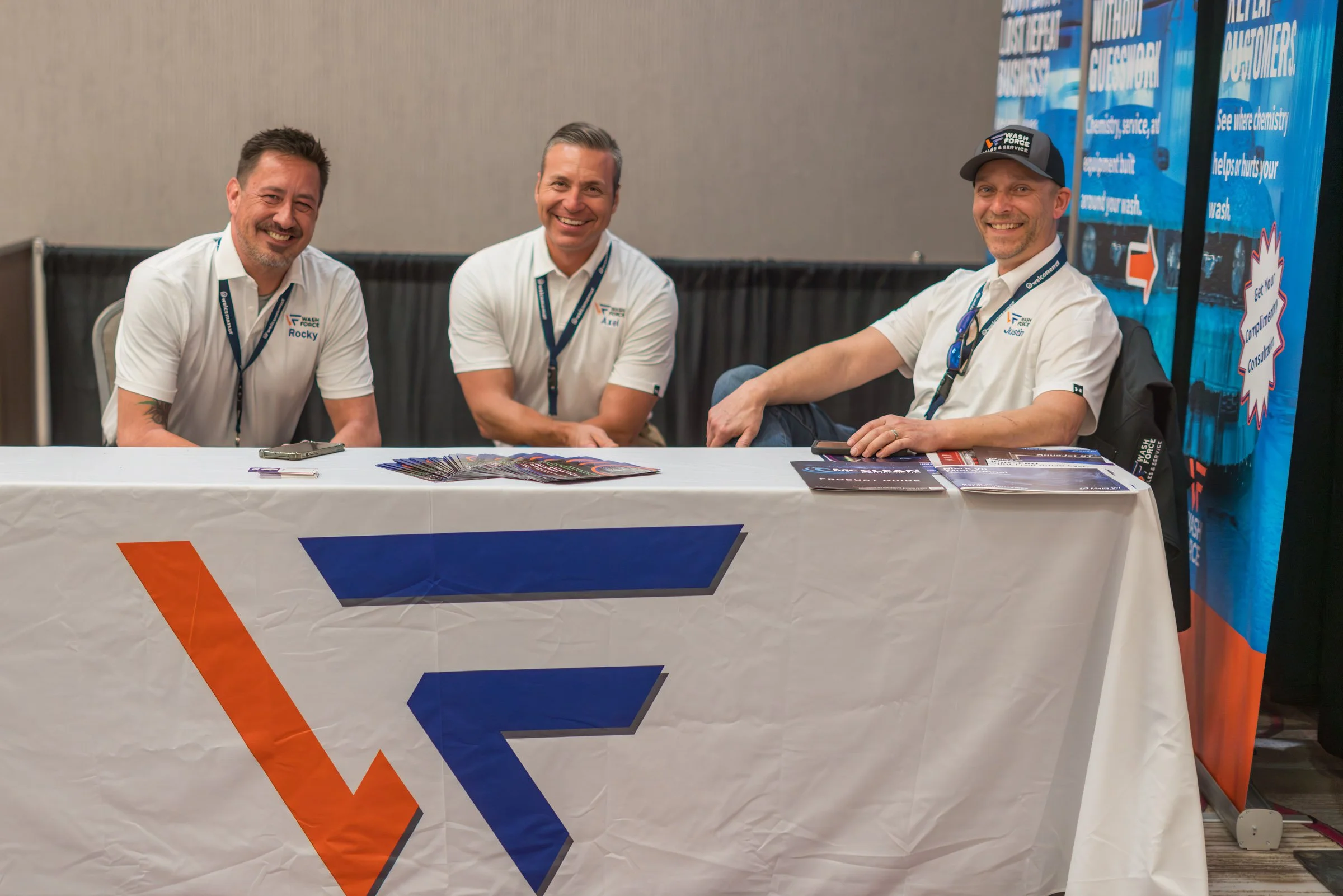 Three men sit behind a table at an indoor event, smiling at the camera. They wear white shirts with company logos and lanyards with badges. Behind them are informational banners with blue backgrounds and text about washing and cleaning services.