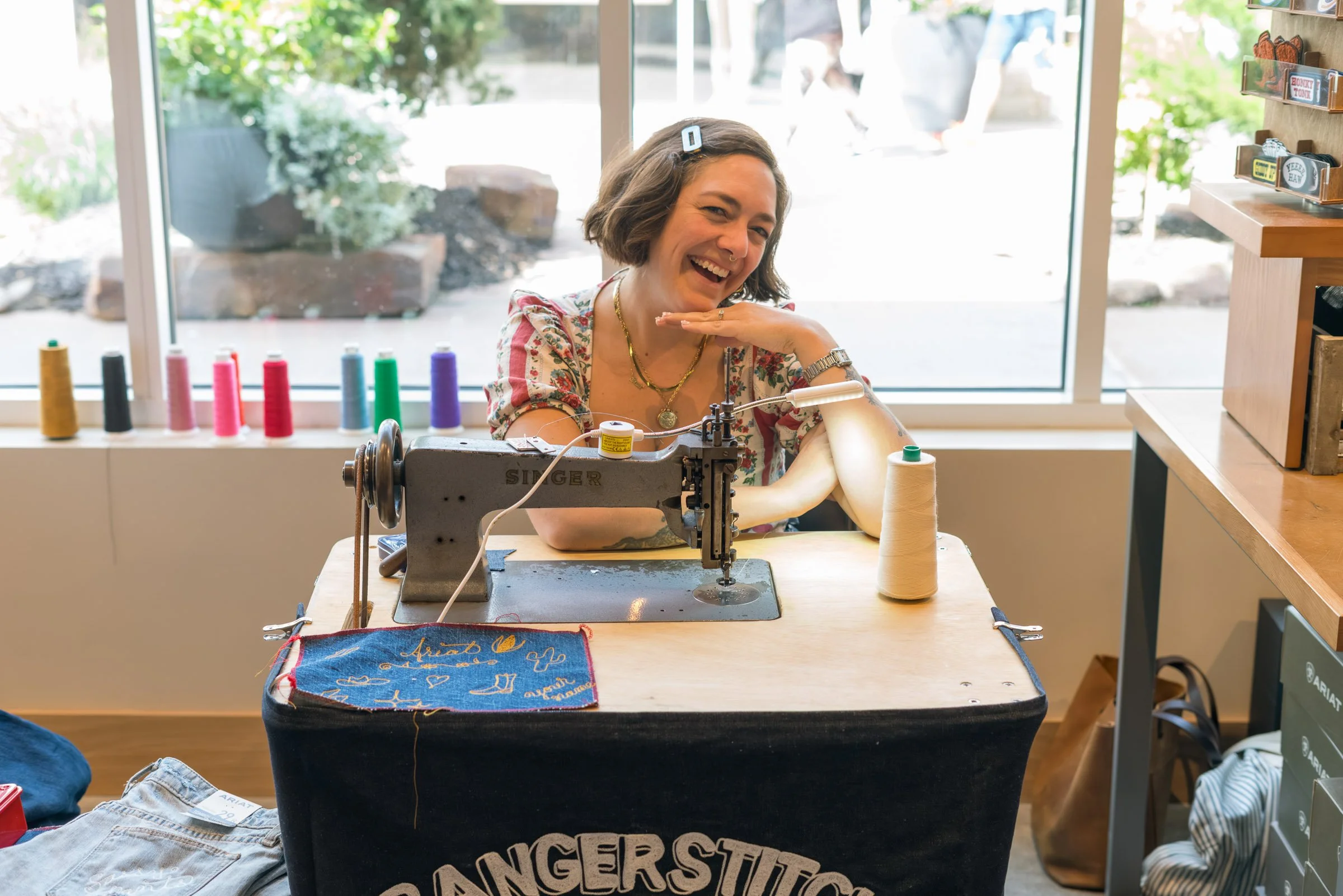 A woman smiling and posing at a sewing machine in a shop, with colorful thread spools on the windowsill behind her and a view outside through a large window.