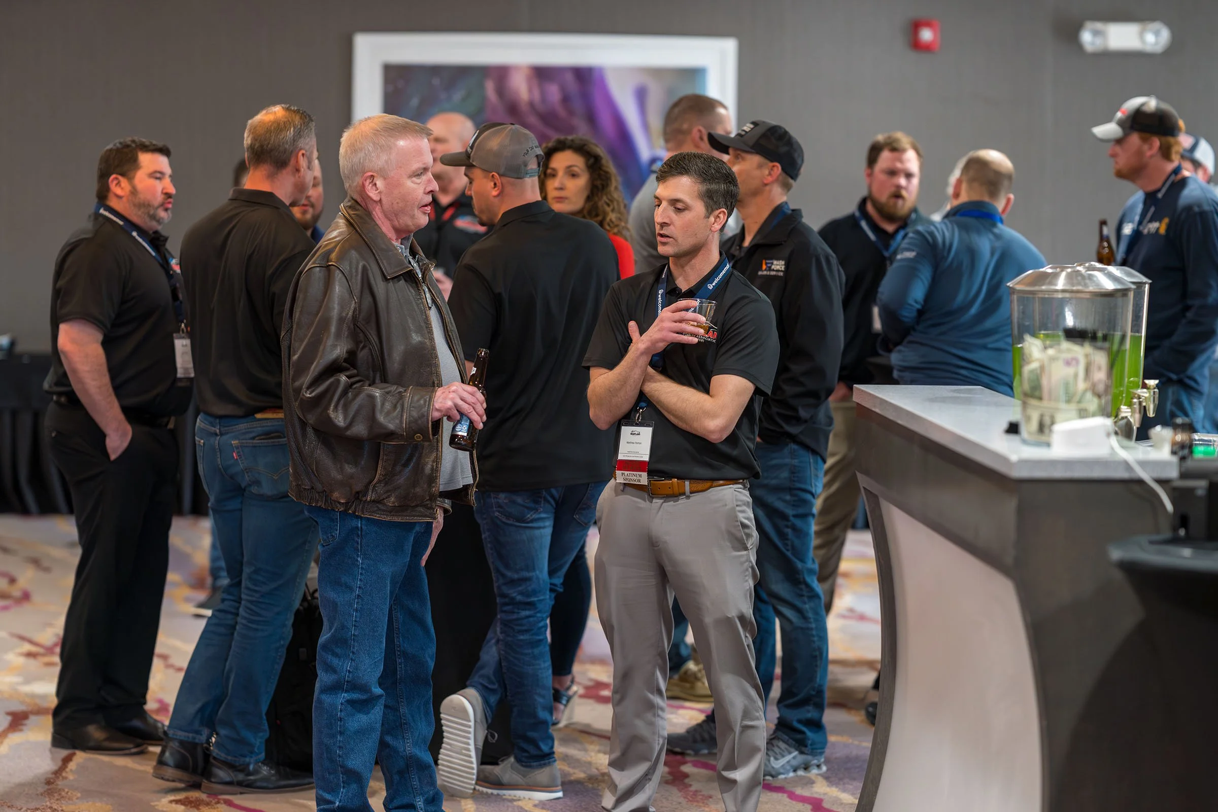 Group of people gathered at a conference or networking event, engaged in conversations, with drinks and a catering station visible.