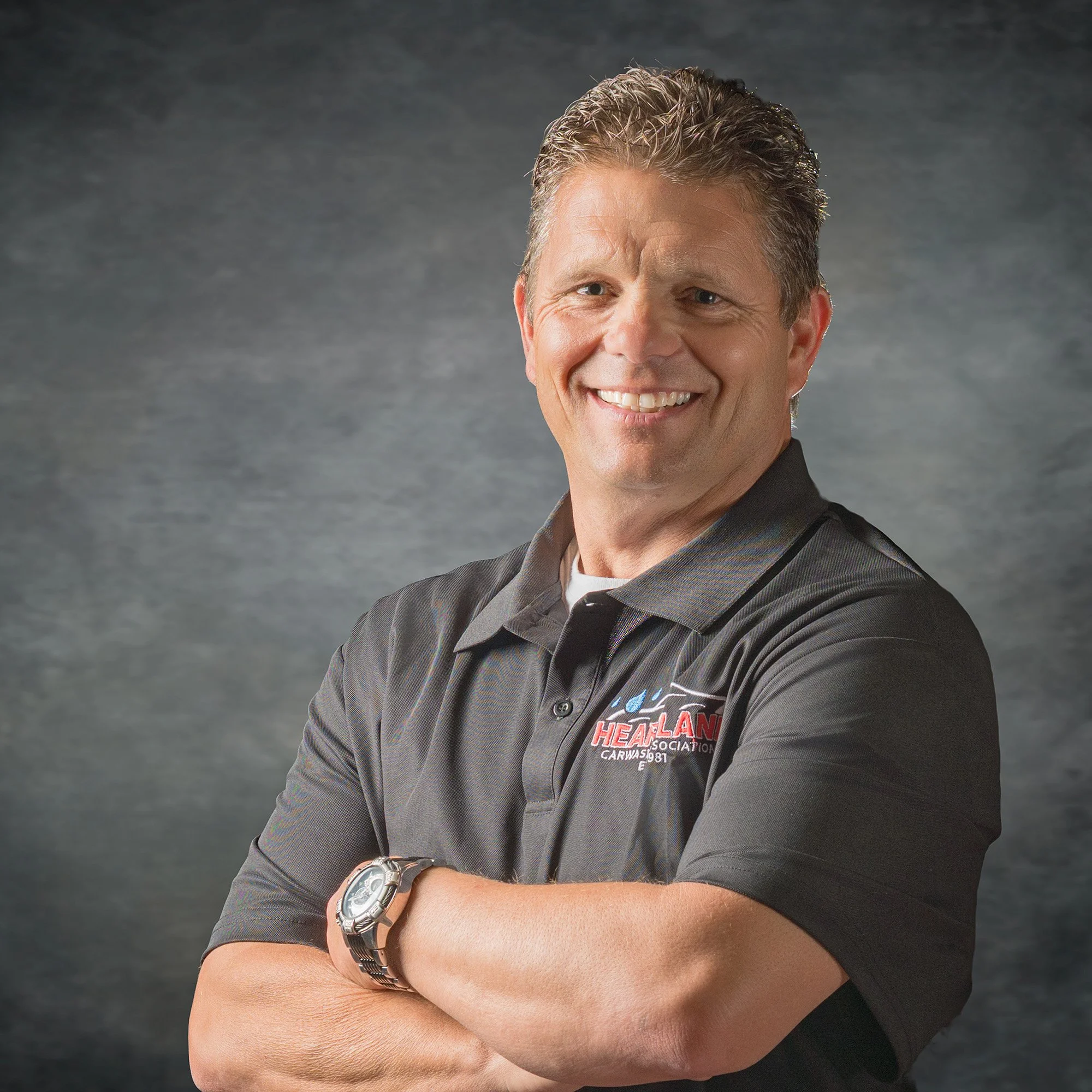 Close-up of a smiling man with gray hair wearing a black collared shirt with logo, a silver watch on his left wrist, against a dark textured background.