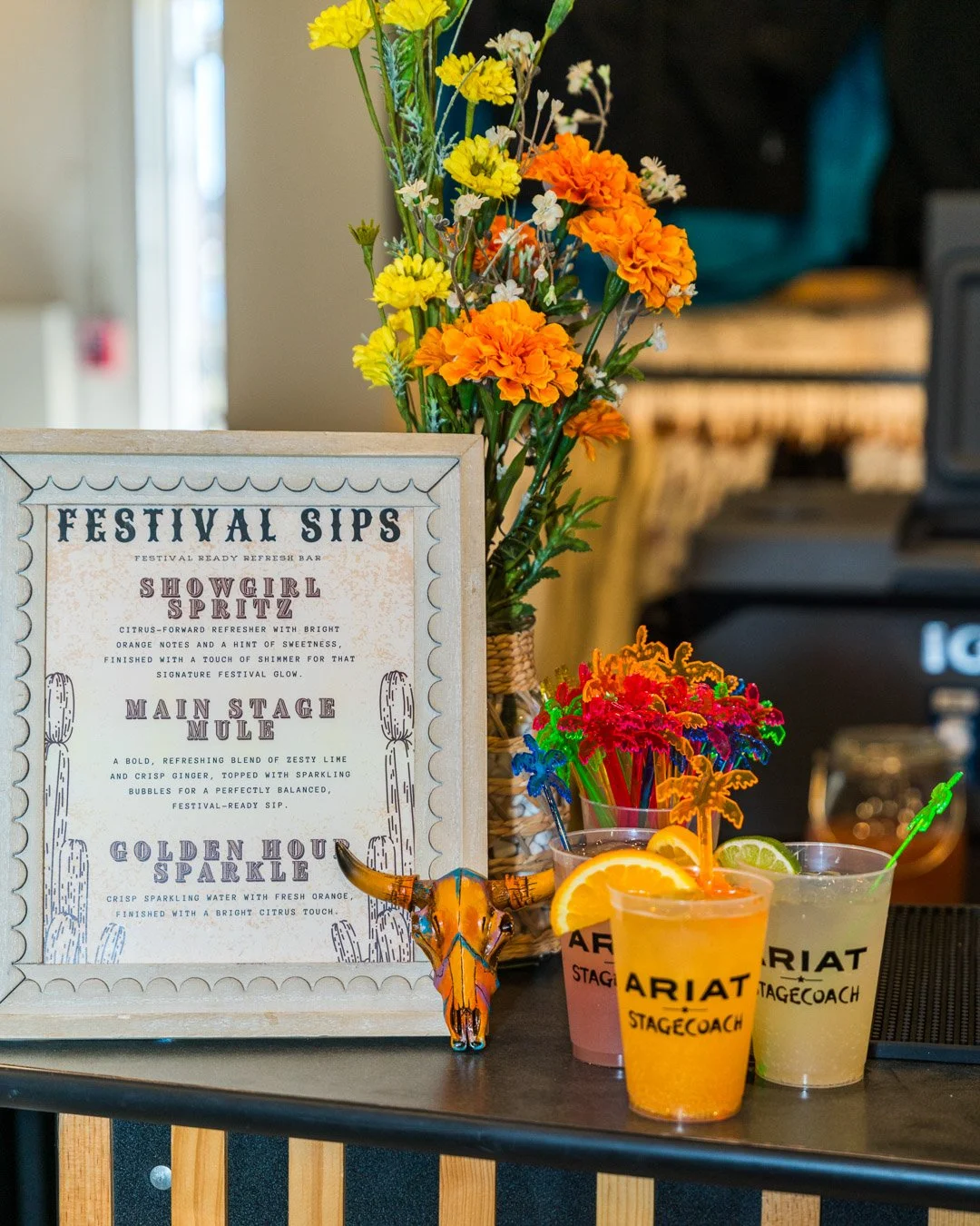Display of colorful festival drinks in front of a floral arrangement and a sign listing festival sips at a bar.