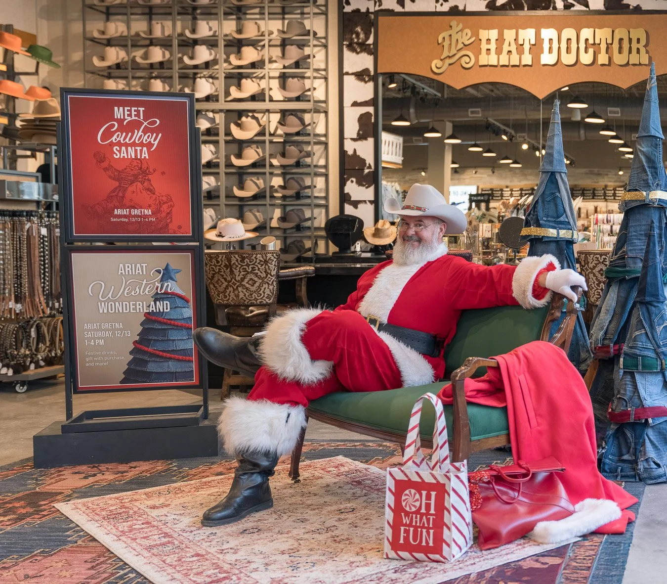 Santa Claus dressed in a red suit with white fur trim, sitting on a vintage green bench in a retail store decorated for Christmas, with signs advertising holiday events for Cowboy Santa and Western Wonderland, and surrounded by cowboy hats, jeans, an