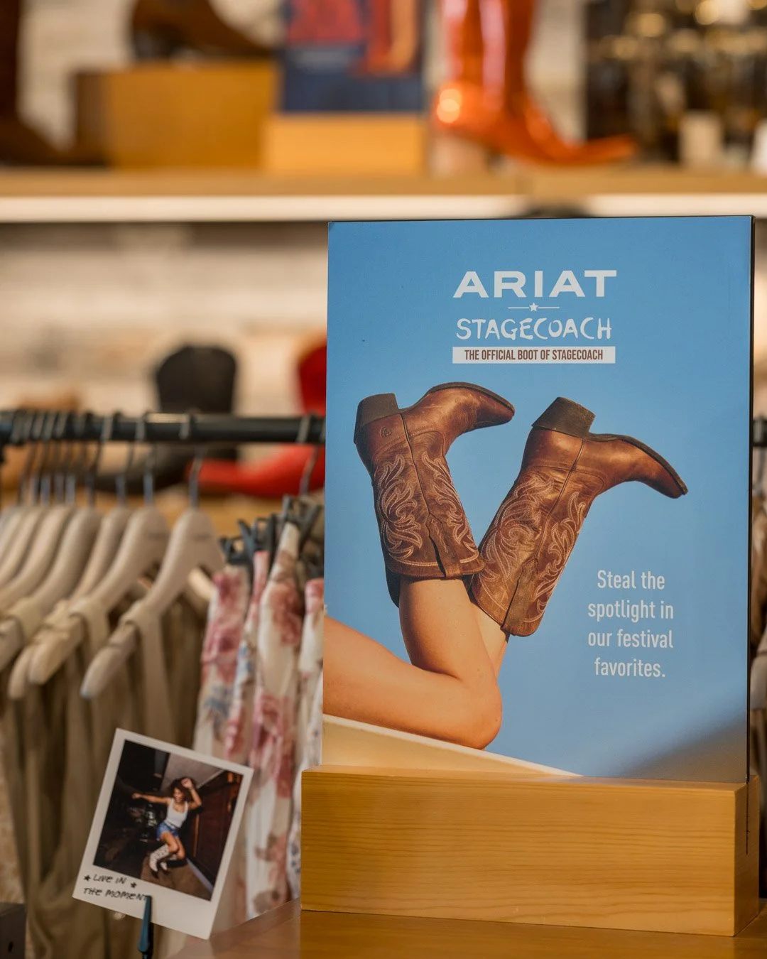 A blue promotional sign for Ariat Stagecoach boots featuring a person with their legs bent at the knees, wearing brown leather cowboy boots with intricate stitching, and the sign saying 'Steal the spotlight in our festival favorites'. There are cloth