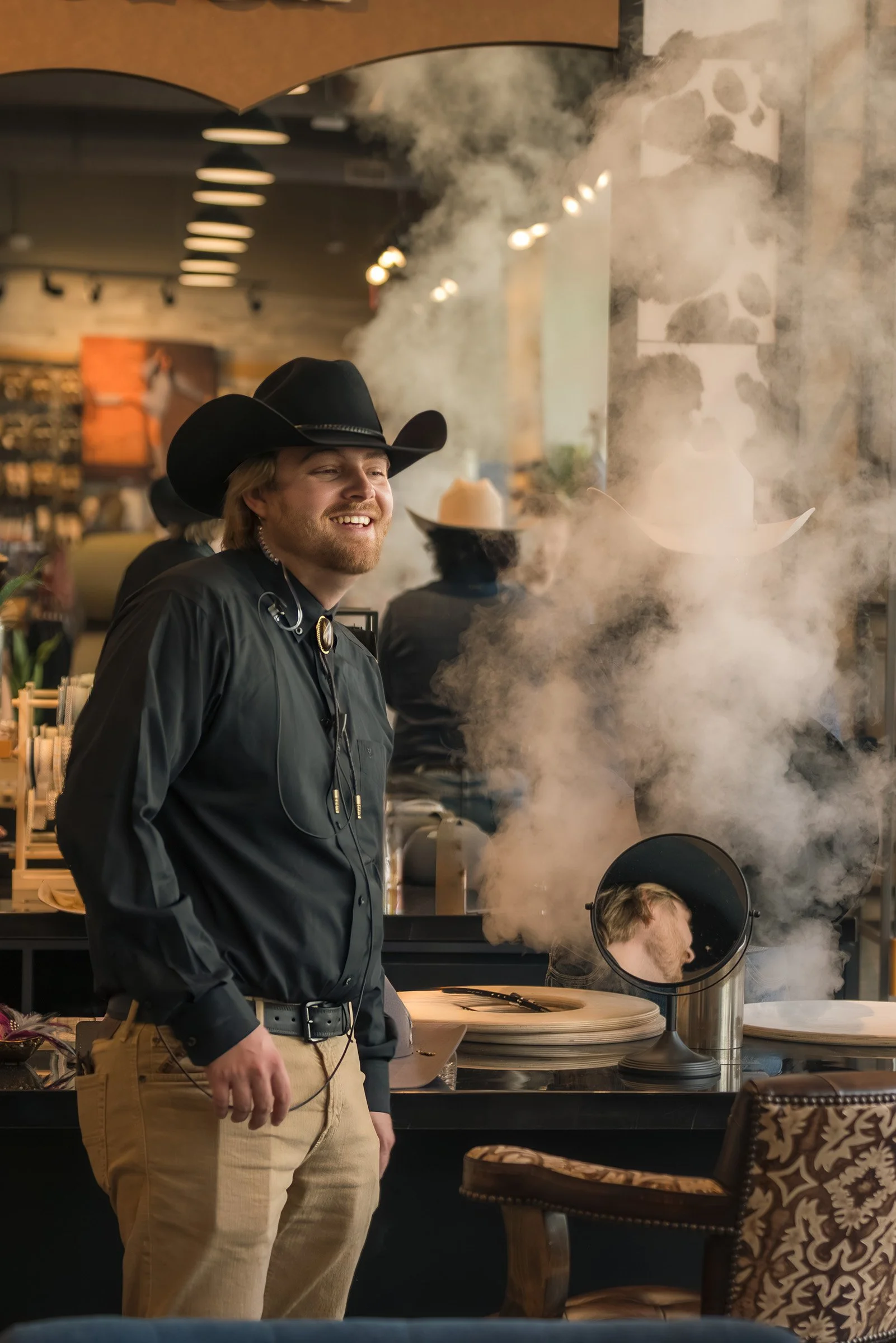 A man wearing a black cowboy hat, black jacket, and beige pants, smiling in a restaurant or cafe with steam or smoke rising from a food warmer on the counter behind him.