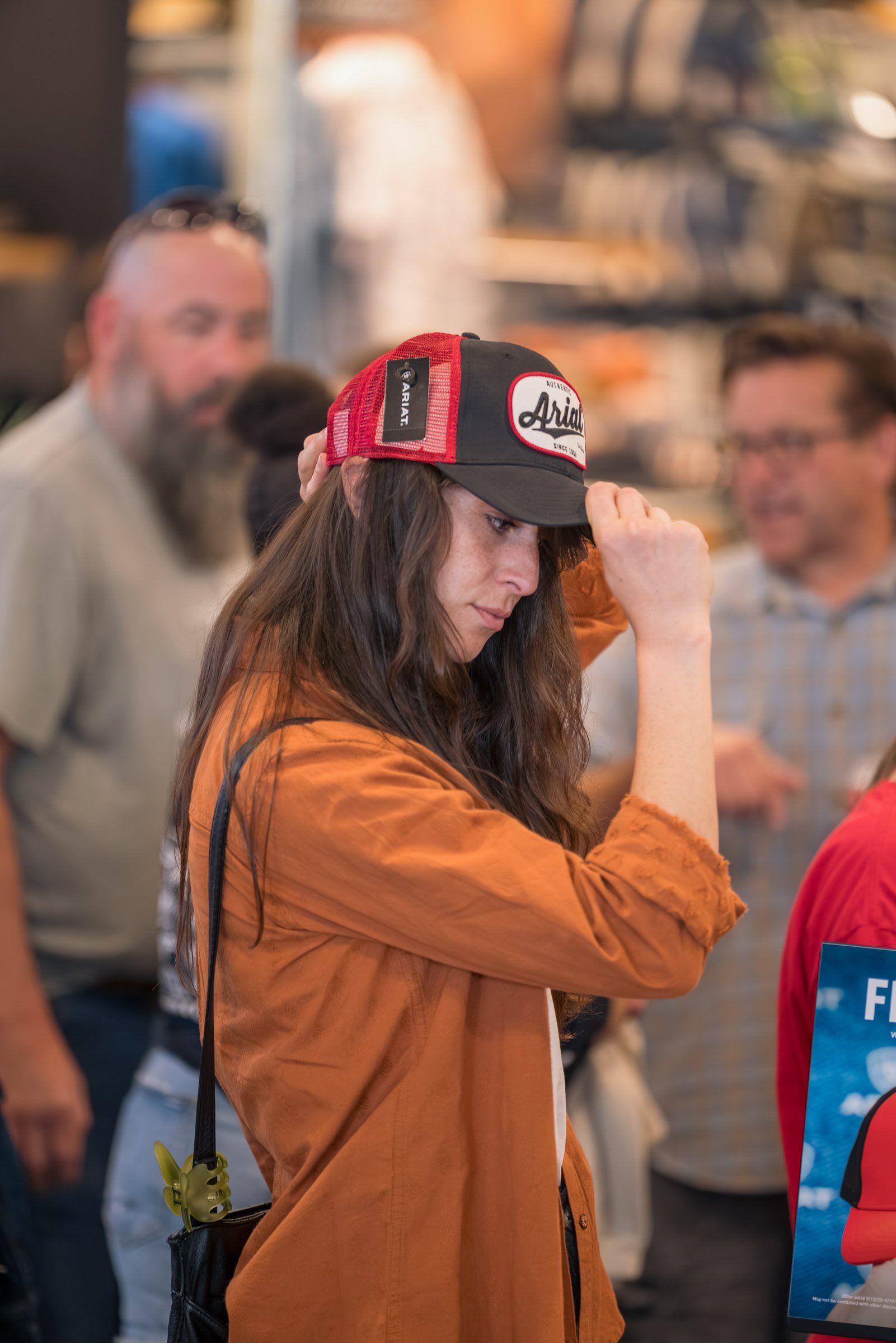A woman with long brown hair wearing an orange jacket and a black and red trucker cap with the word 'Ariat' on the front, adjusting her cap indoors at a crowded event.