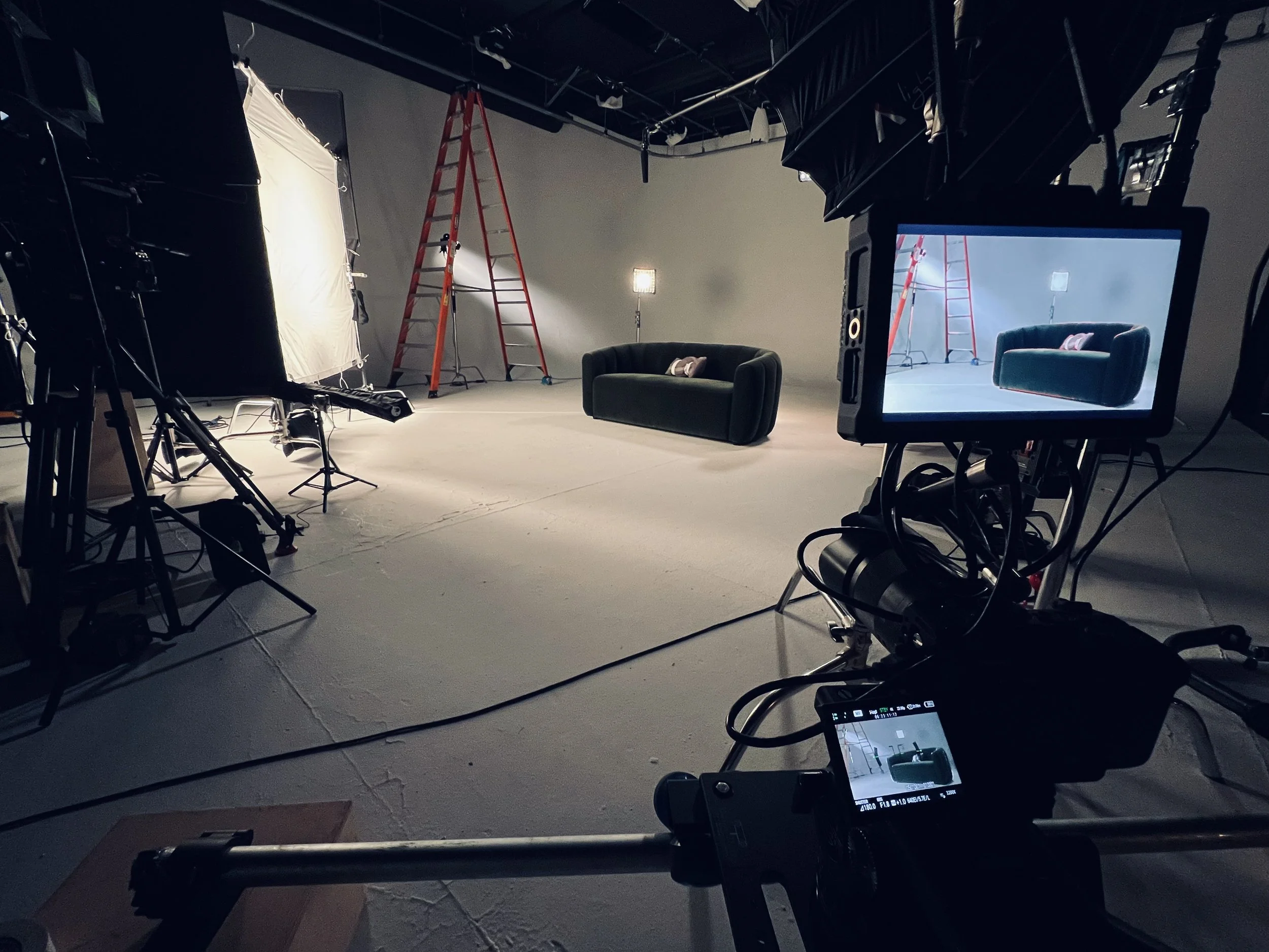 Empty video production studio with two black couches, a red ladder, lighting equipment, and a camera monitor showing the scene setup.