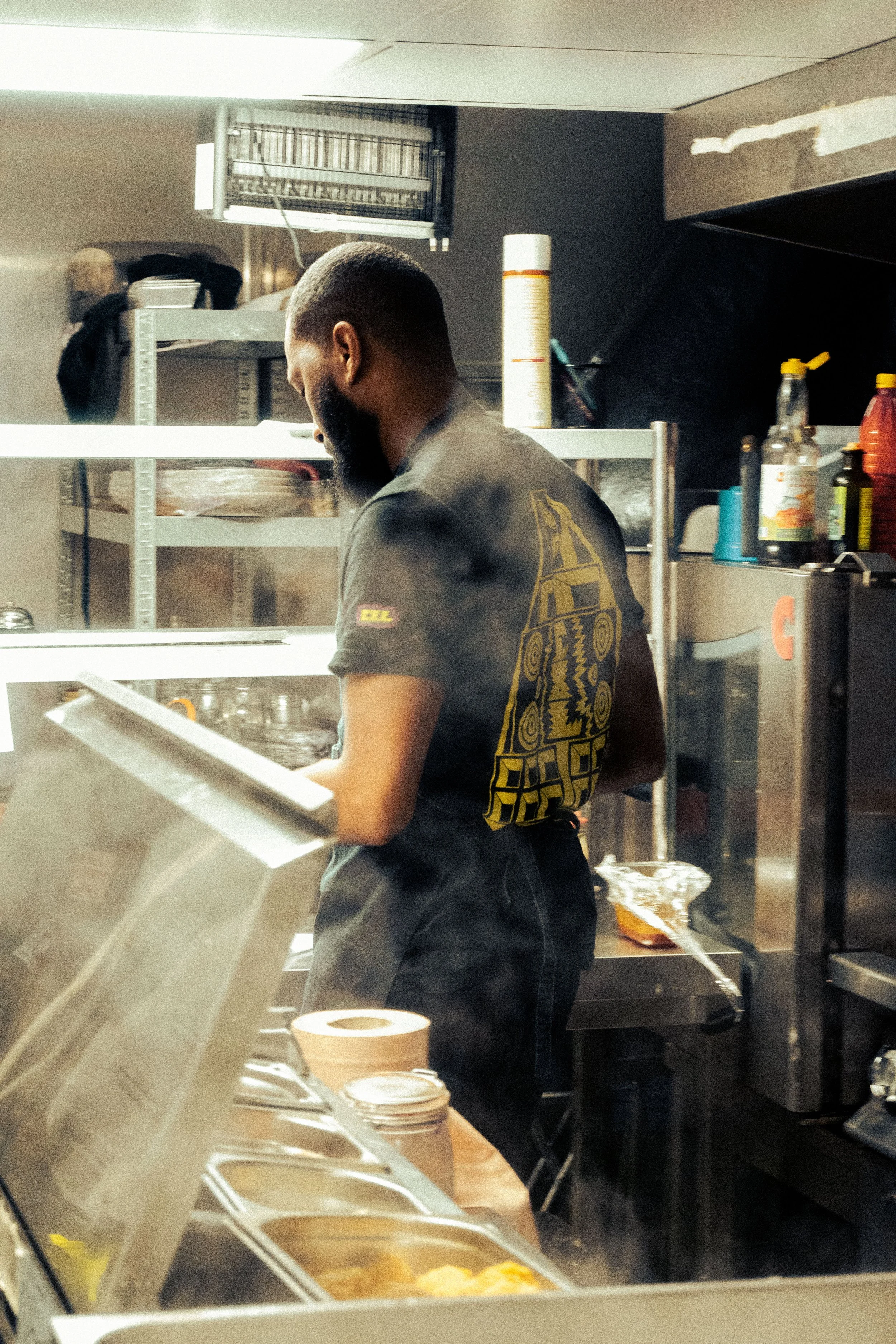 Photo stylisée d'un cuisinier vue de profil dans une cuisine de restaurant. Il porte un t-shirt noir avec un design Bouyon sur le dos et travaille derrière un comptoir avec divers ingrédients et ustensiles.