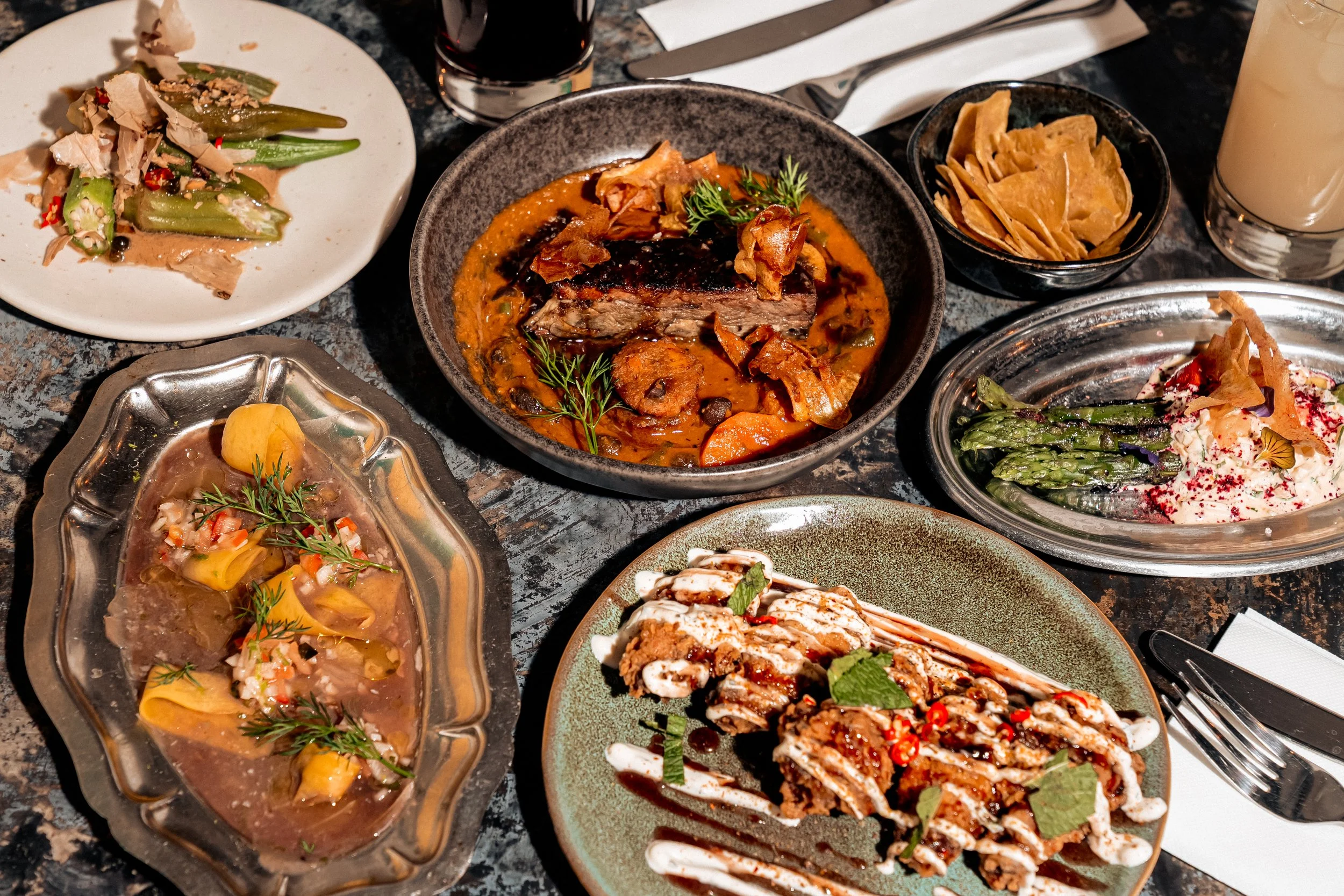 Assortiment de plats africains et cocktails avec différents plats: mafé, gombos, ceviche, fried chicken, plantain, asperges sur une table en acier design - cuisine Nwaar afro-caribéenne Belleville Paris