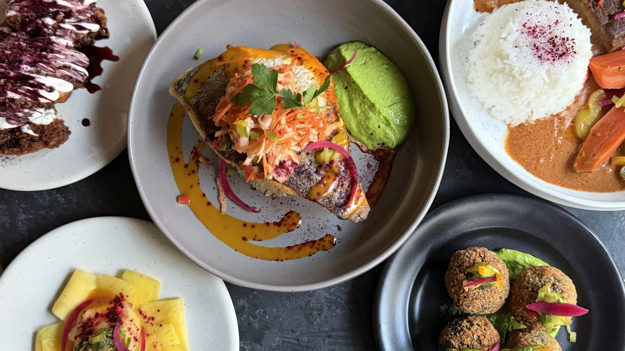Assortiment de plats africains et caribéens, incluant poisson grillé, riz avec sauce et légumes, croquettes arancini, accompagnés de diverses sauces et garnitures.