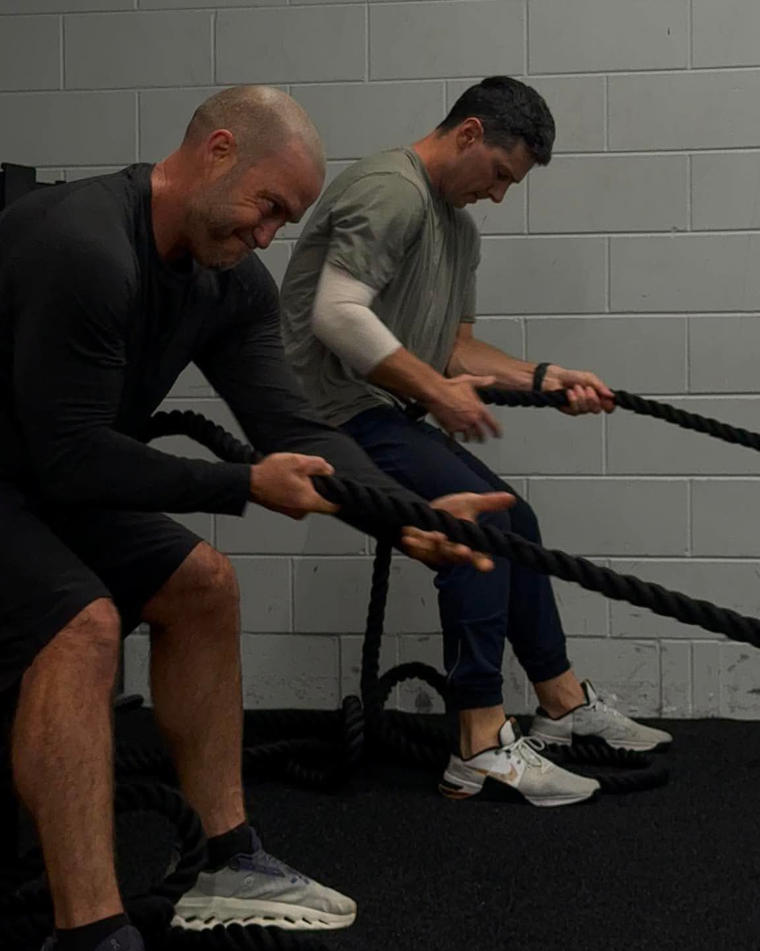 Two men engaging in a battle rope workout in a gym with gray cinder block wall.