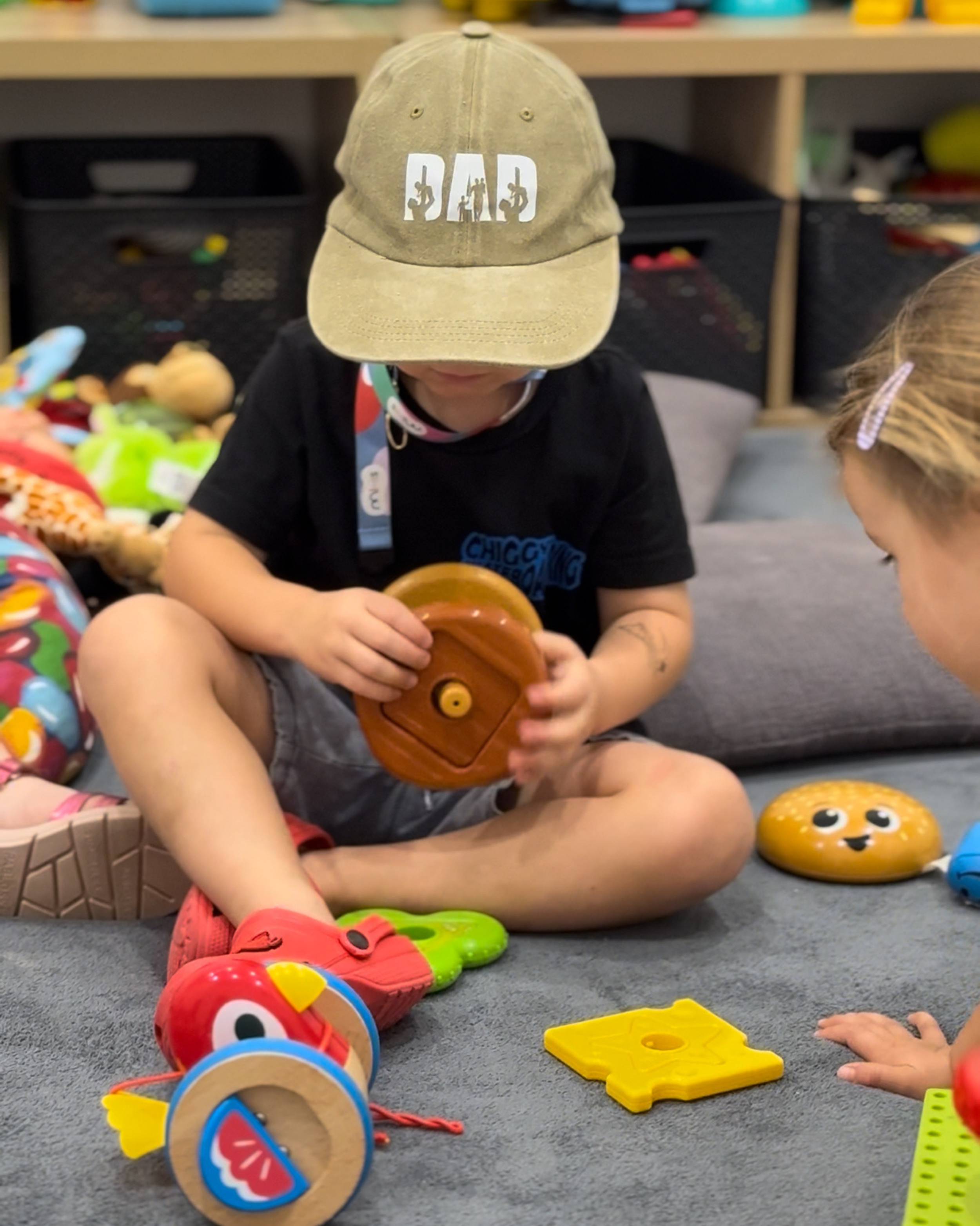 A child wearing a khaki cap, playing with colourful toys on a gray carpeted floor in a room with a shelf in the background.