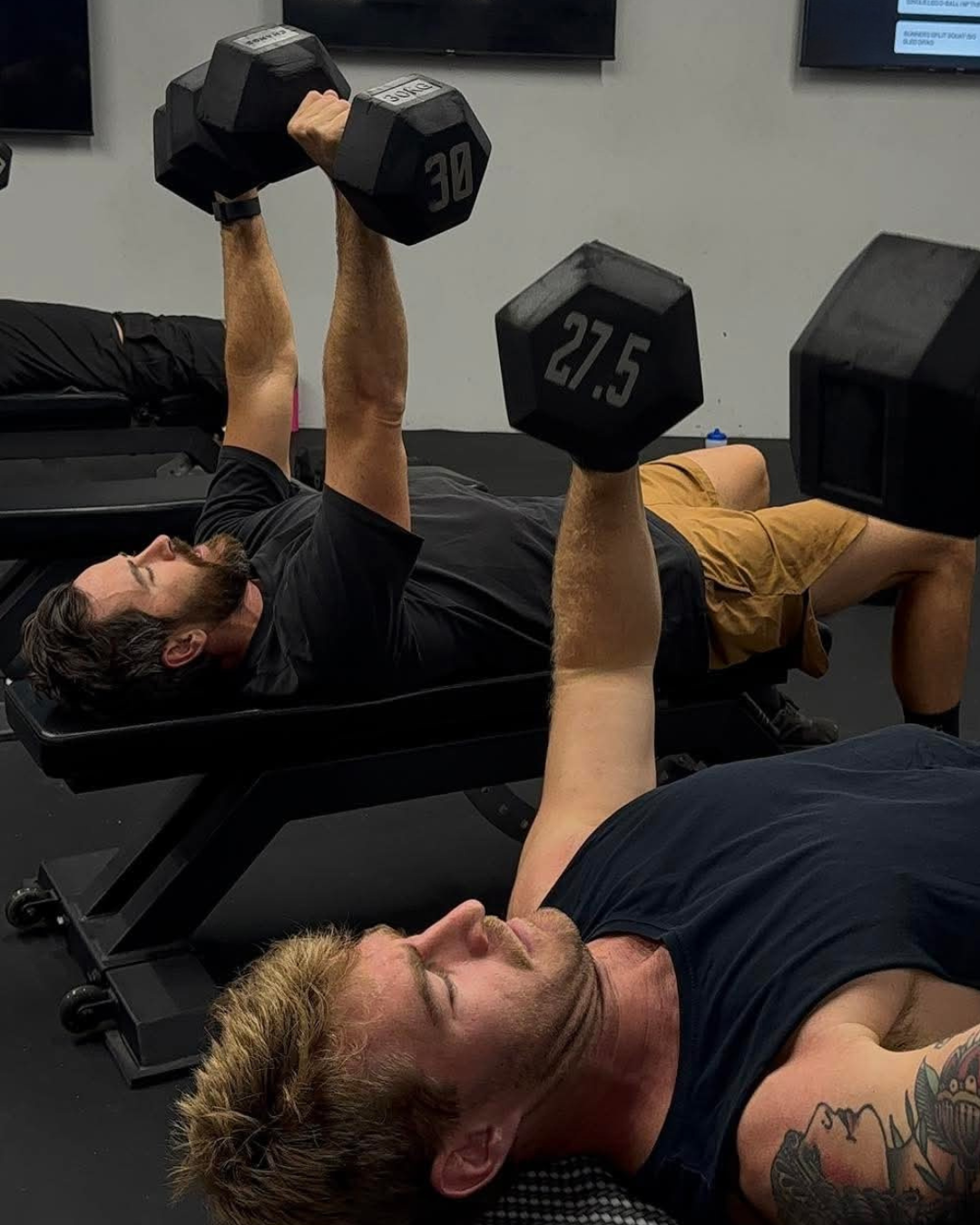 Two men are lying on exercise mats, lifting dumbbells with their arms extended upward, in a gym setting. One man has a tattoo on his arm, both appear focused during their workout.
