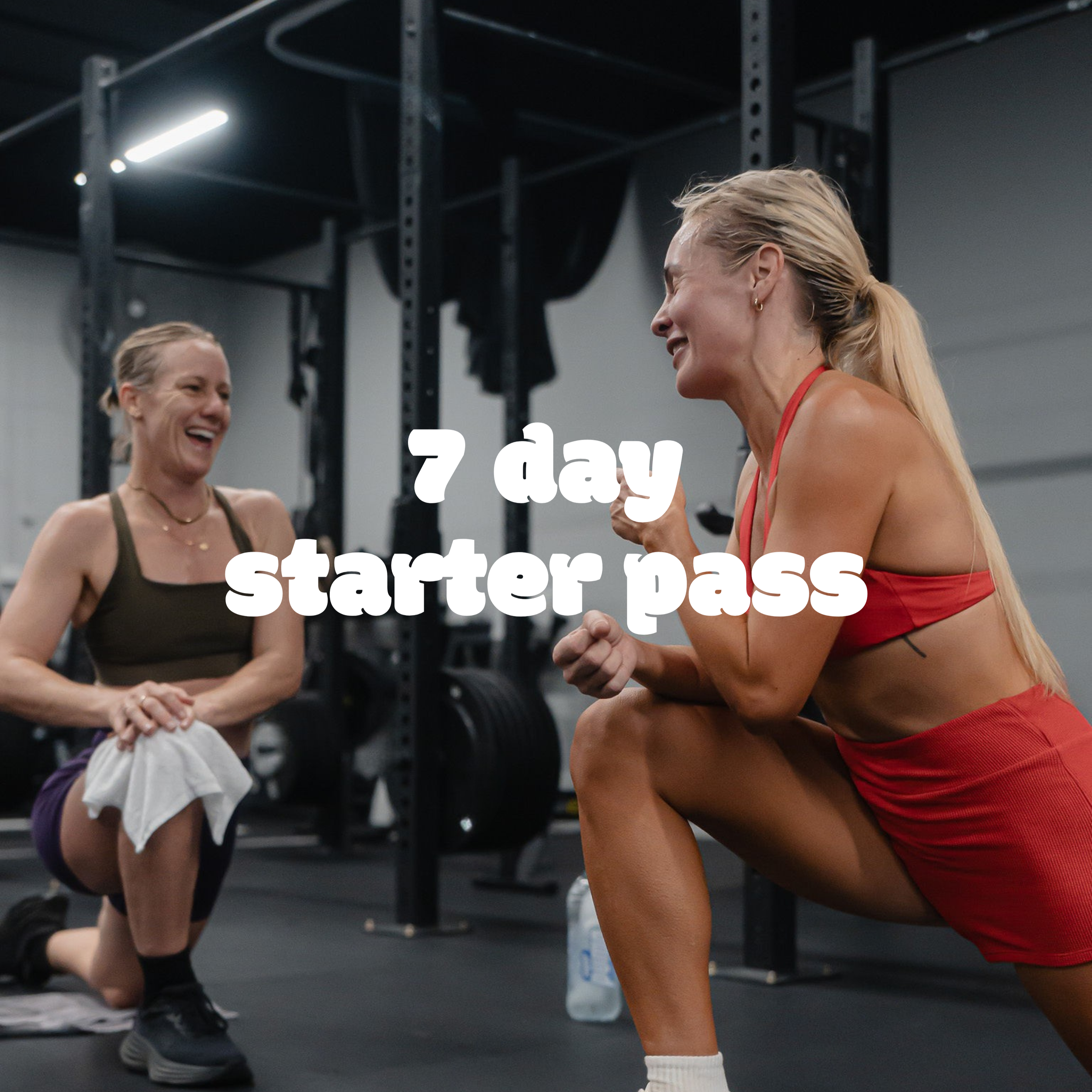 Two women are smiling and laughing while engaging in a workout or fitness session in a gym. One woman is wearing a black tank top and the other is wearing a red sports bra and red shorts. They are in a crossfit or gym setting, with black metal workout equipment in the background.