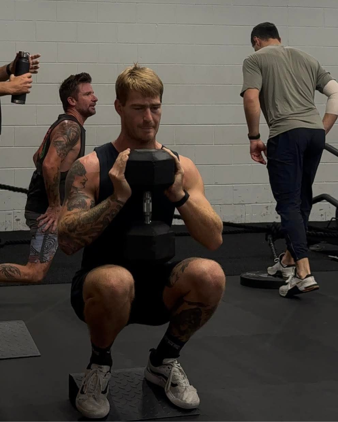 A man squatting on a platform in a gym while holding a dumbbell at shoulder height, focusing on his exercise. In the background, two other men are present, one kneeling on the floor and another with his back turned, all surrounded by gym equipment and a gray brick wall.