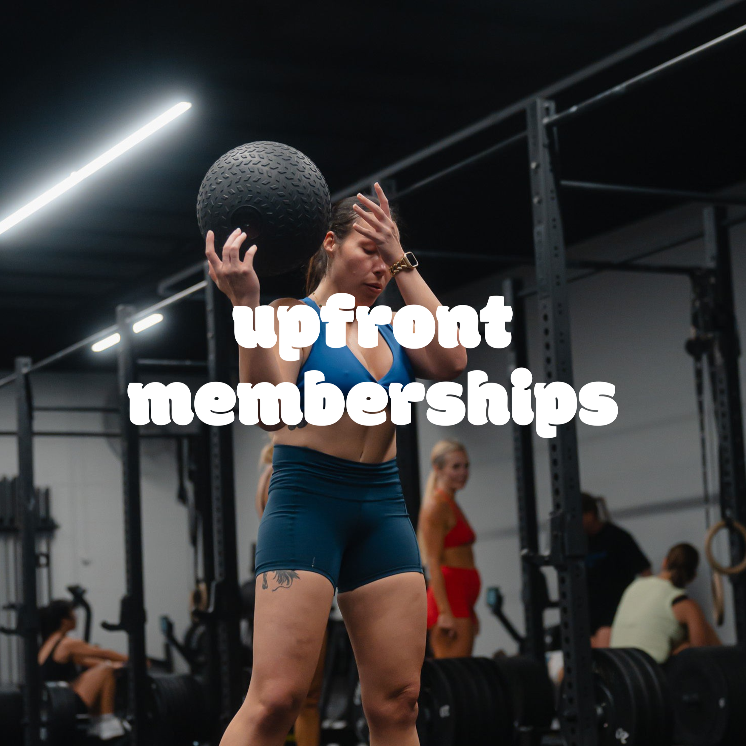 A woman in athletic attire throwing a medicine ball over her shoulder in a gym setting with other women in the background.