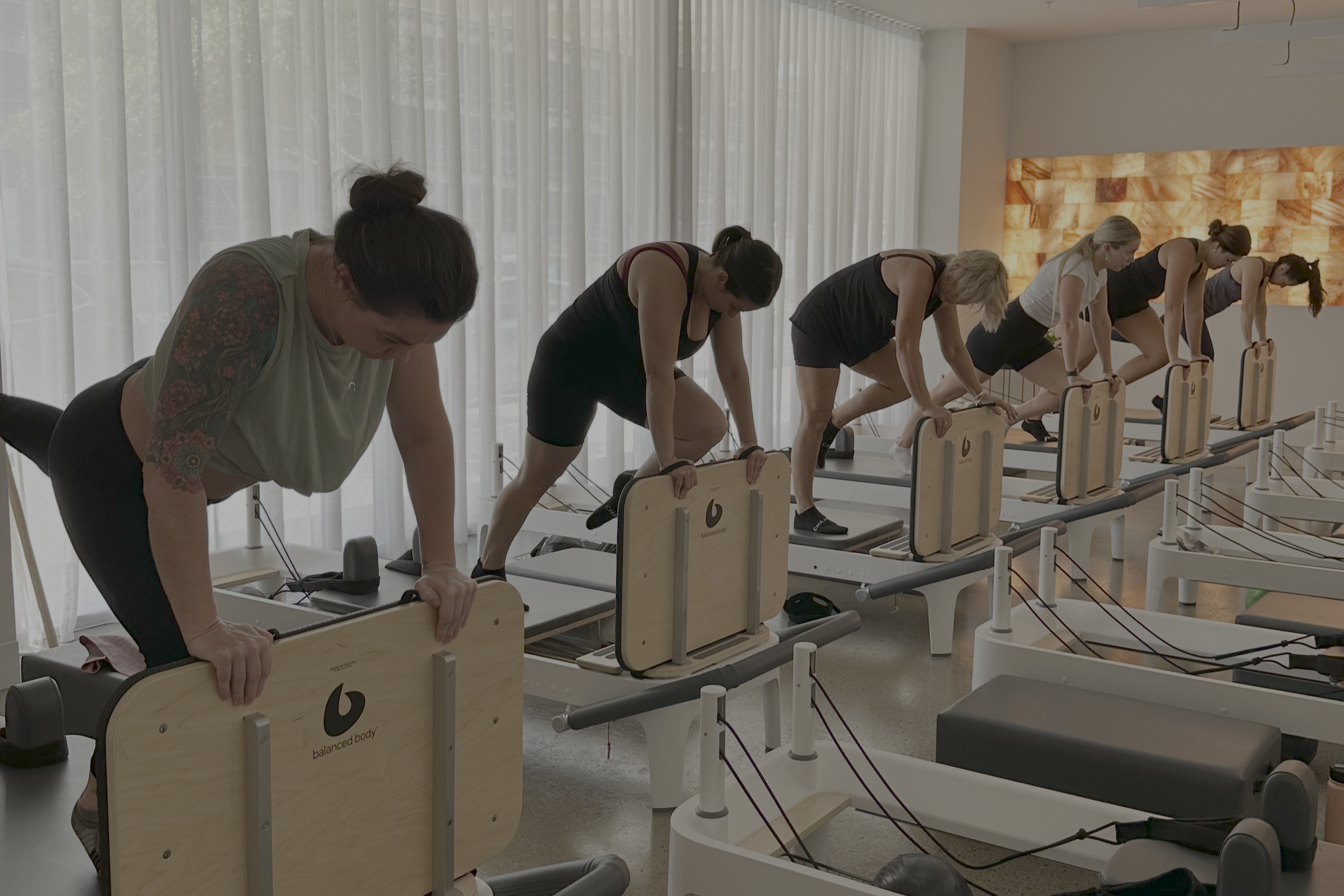 Several women participating in a Pilates reformer class in a bright studio with large windows and sheer curtains.