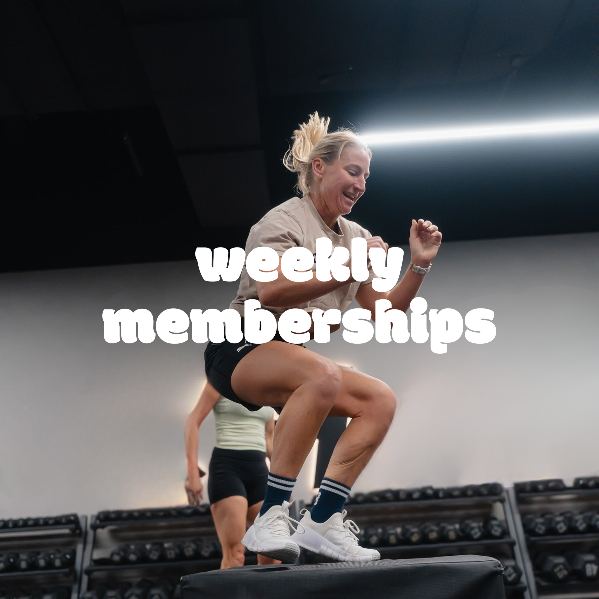 Woman smiling and jumping on plyo box at the gym, with weights in the background, wearing white sneakers, striped socks, and athletic clothes.