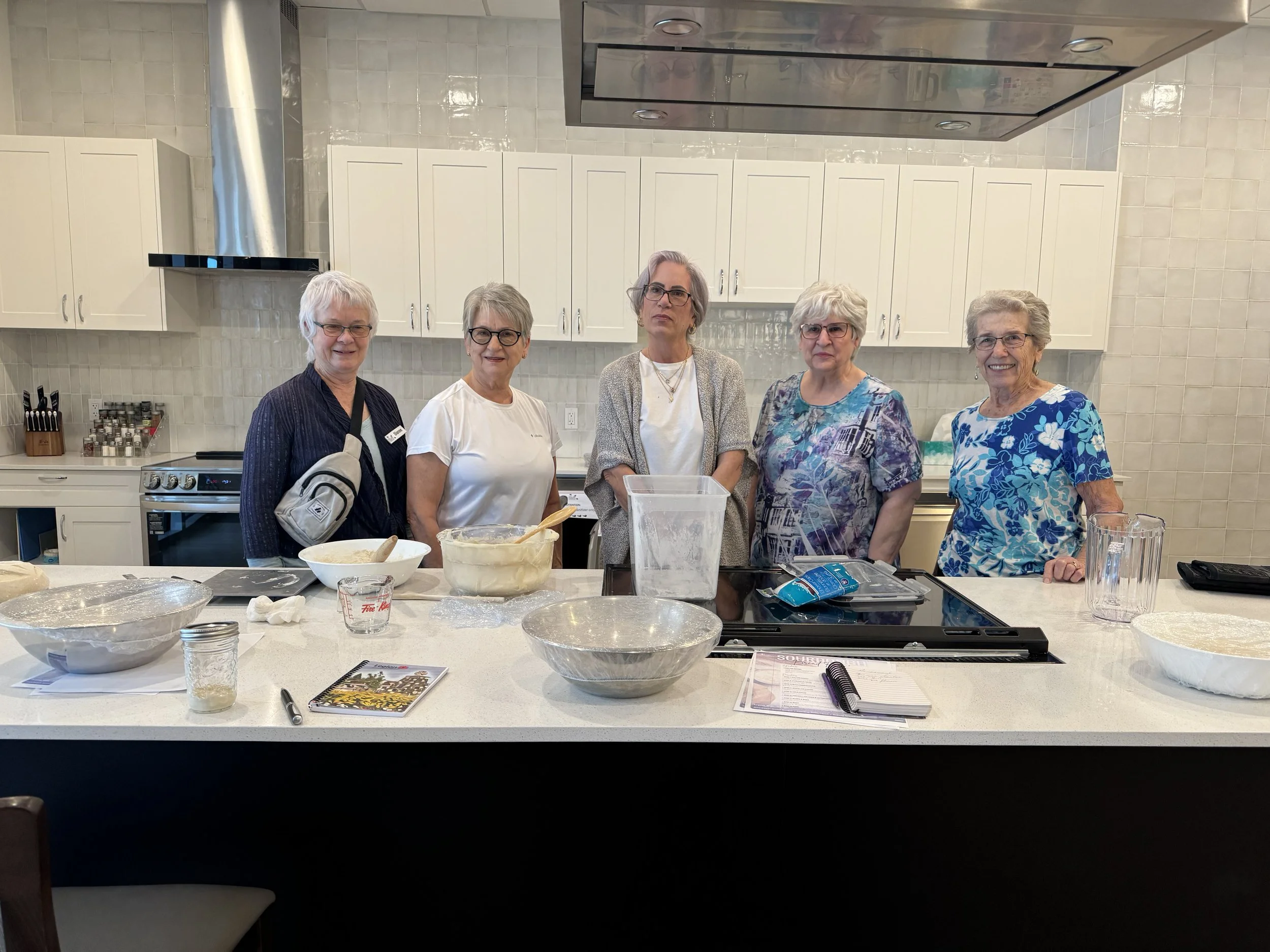 Six women standing behind a kitchen counter with baking supplies, smiling and preparing for baking.