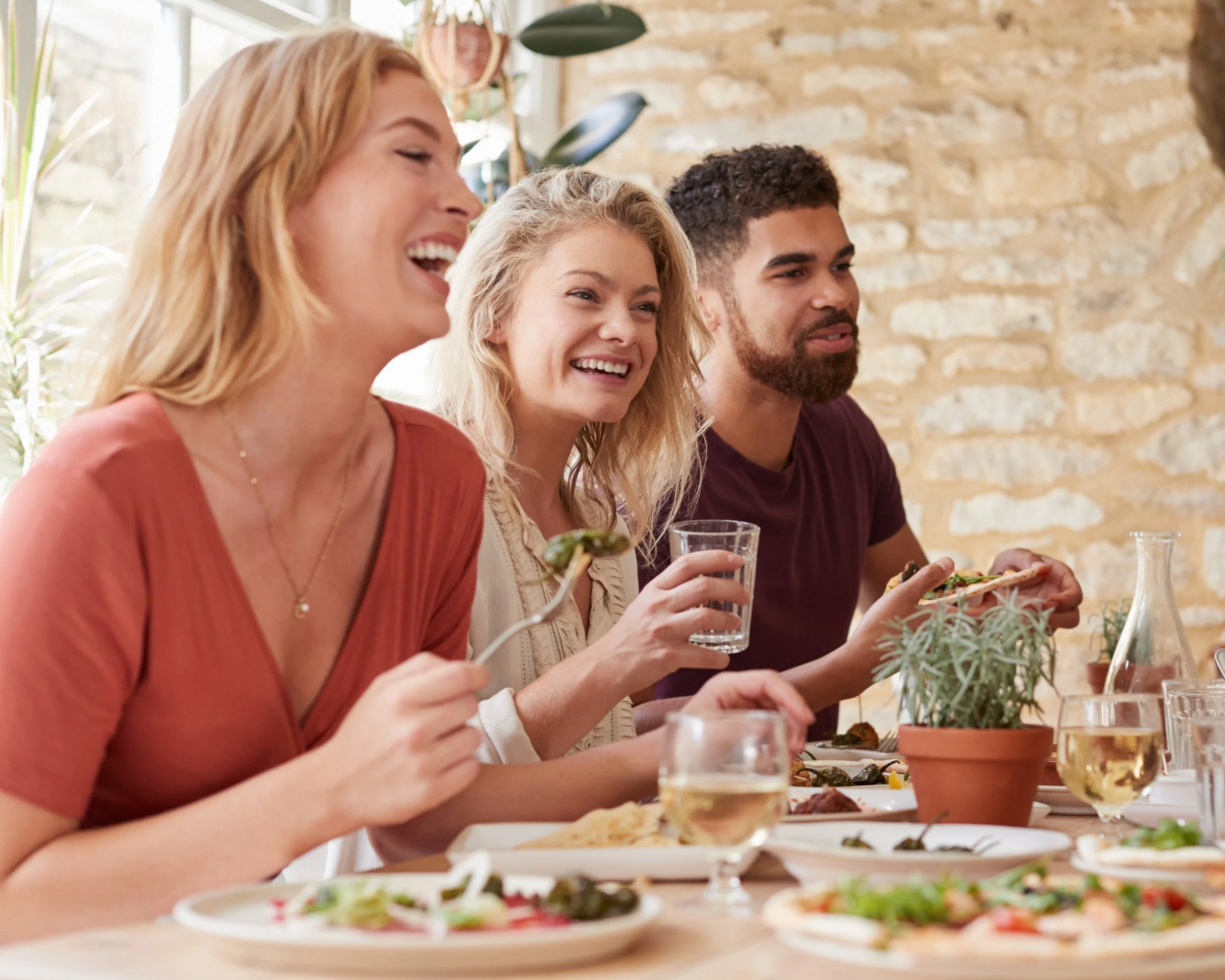 Three friends enjoying a meal together at a restaurant, smiling and laughing. They are sitting at a table with plates of food, glasses of white wine and water, and a potted plant as a centerpiece, in a cozy indoor setting with exposed brick walls.