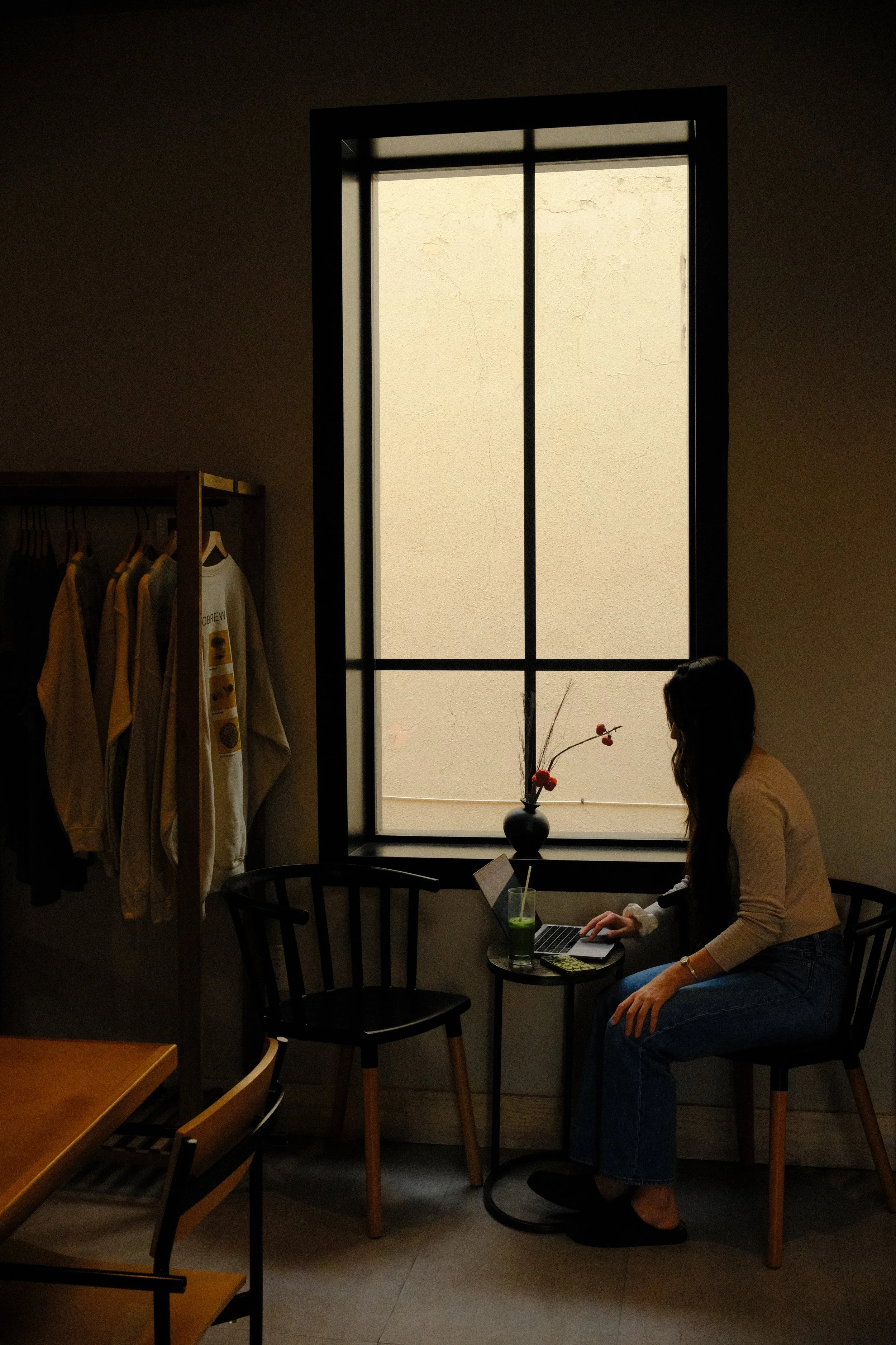 Woman in a cream sweater and jeans working on a laptop at a small café table by a sunlit window, with a fresh matcha latte beside her - Hello Holistic Studio.