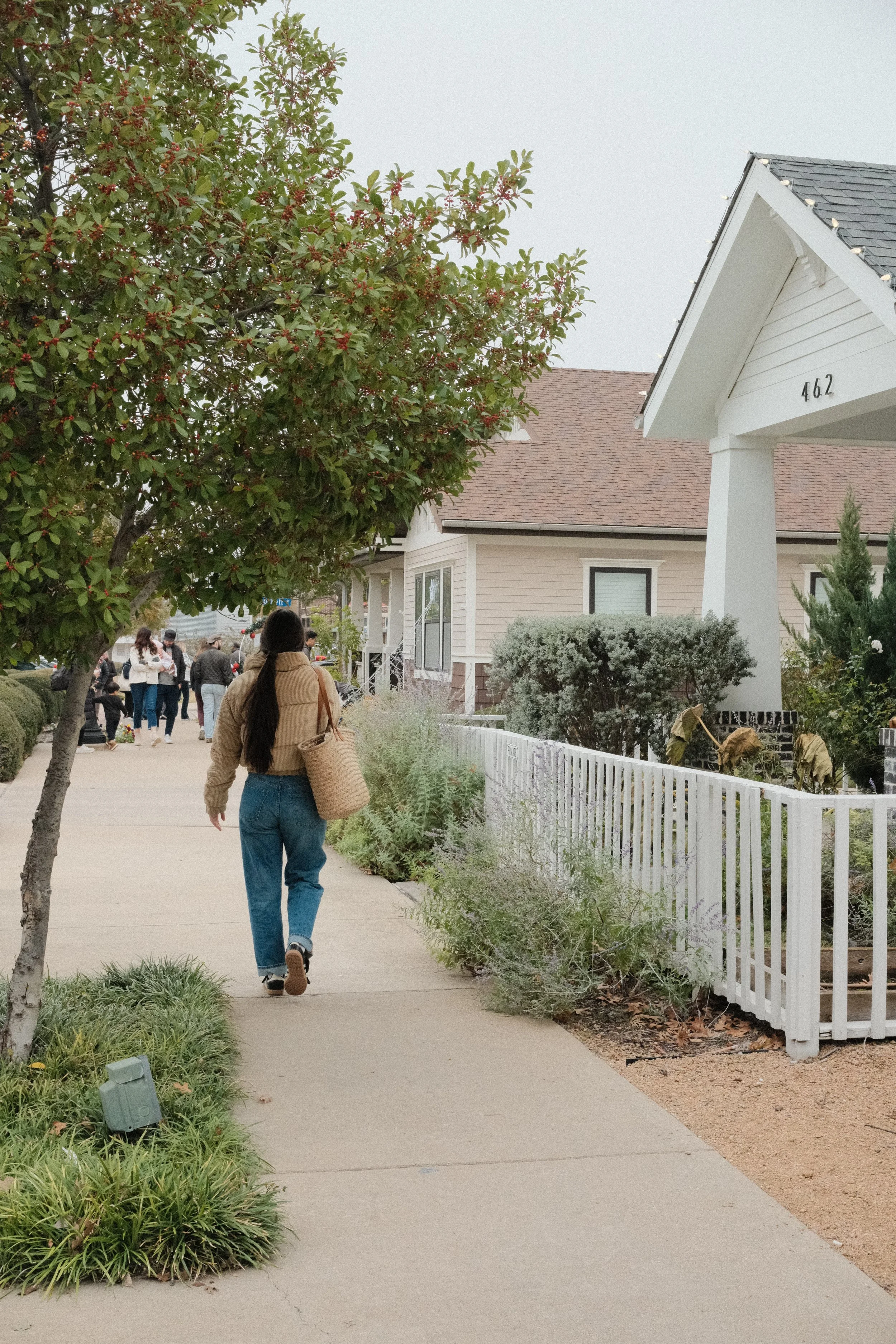 Woman in a tan corduroy puffer jacket and jeans walking down a tree-lined sidewalk past a white picket fence and craftsman-style home, carrying a woven basket - Hello Holistic Studio.