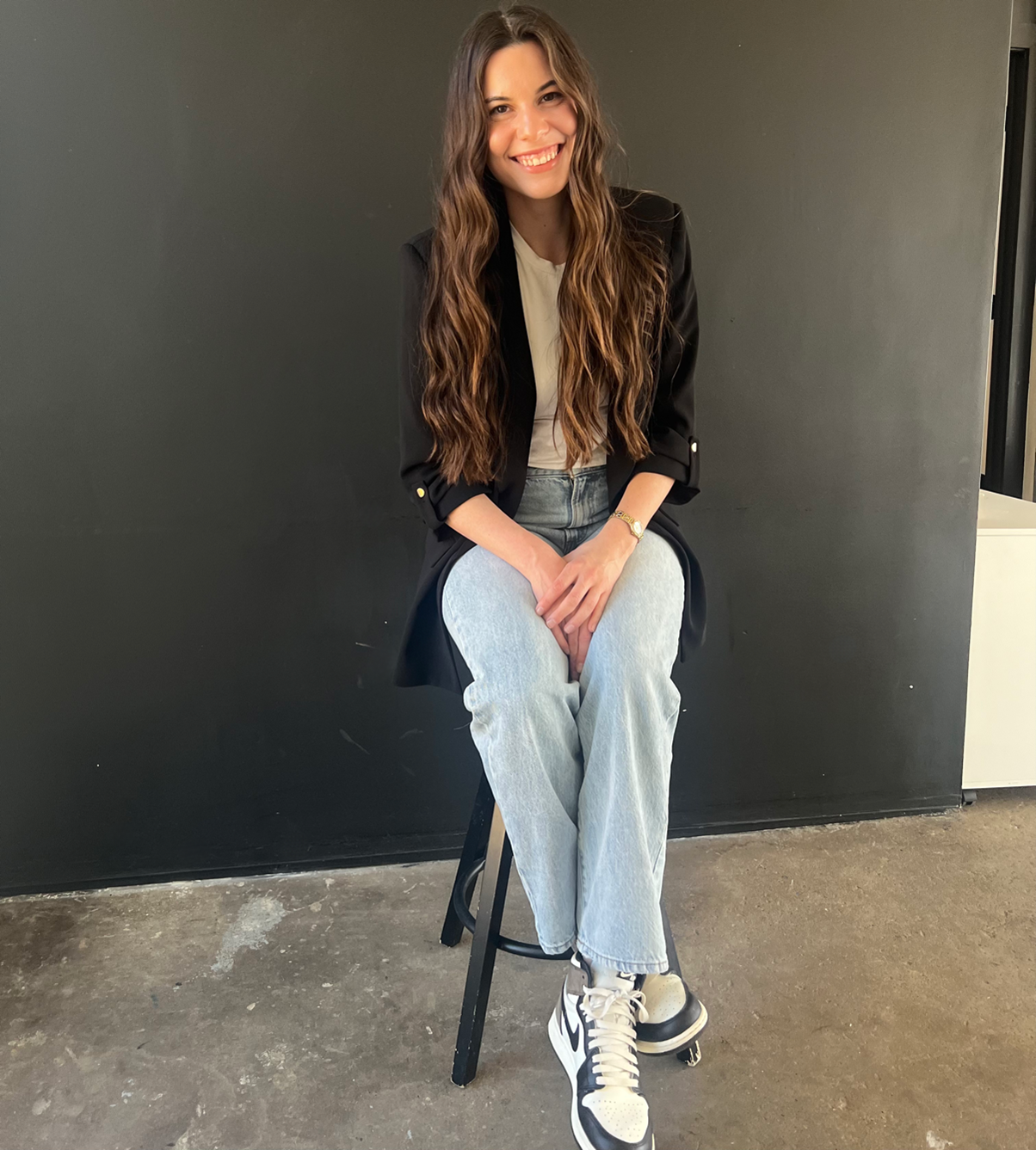Young woman with long wavy hair smiling and sitting on a black stool against a black wall, wearing a black blazer, white shirt, light wash jeans, and sneakers.
